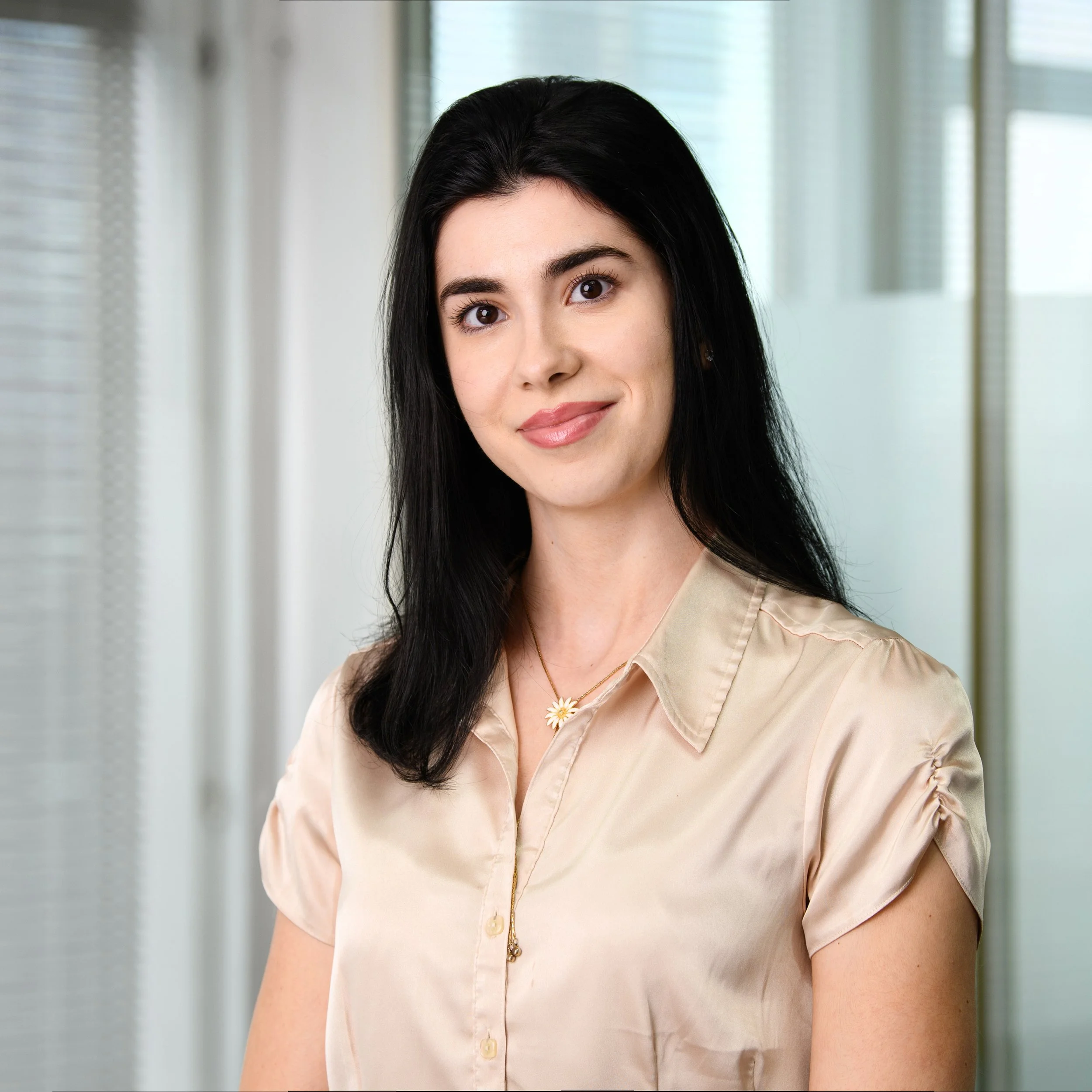 Close-up of a young woman with long black hair, wearing a beige button-up shirt and a daisy-shaped necklace, standing in front of a light-colored, windowed background.