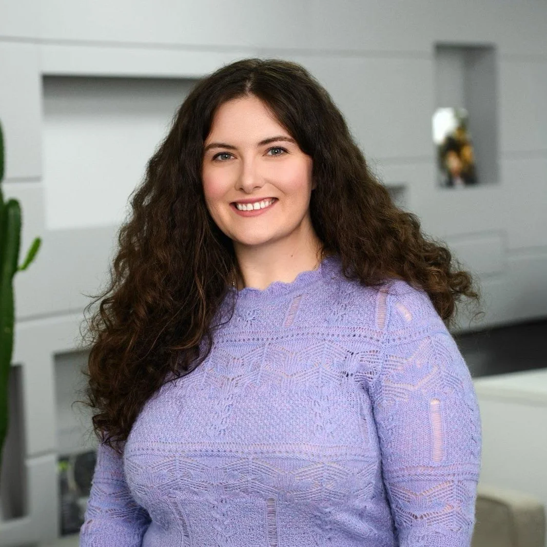 A woman with long, curly dark brown hair wearing a lavender lace-knit sweater, smiling indoors with a white wall, a cactus, and a built-in shelf in the background.