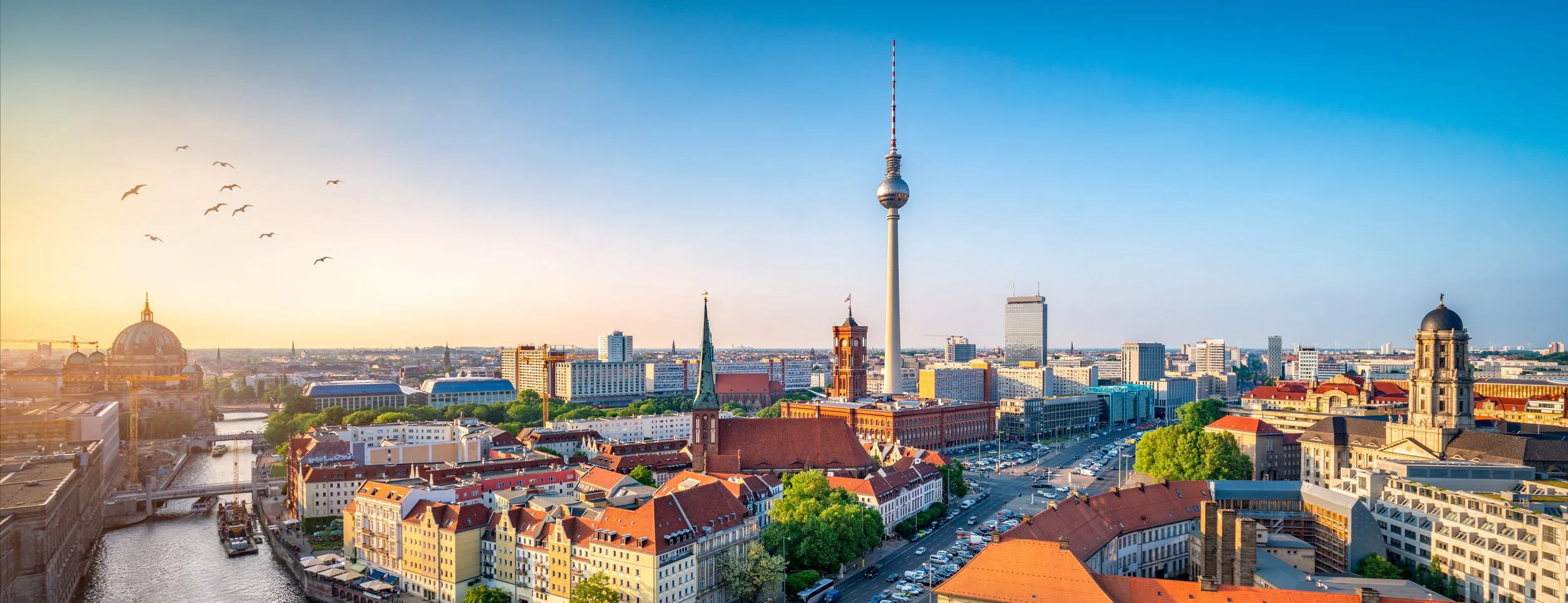 A cityscape of Berlin, Germany at sunset, featuring the Berlin TV Tower (Fernsehturm) and historic buildings along the Spree River.
