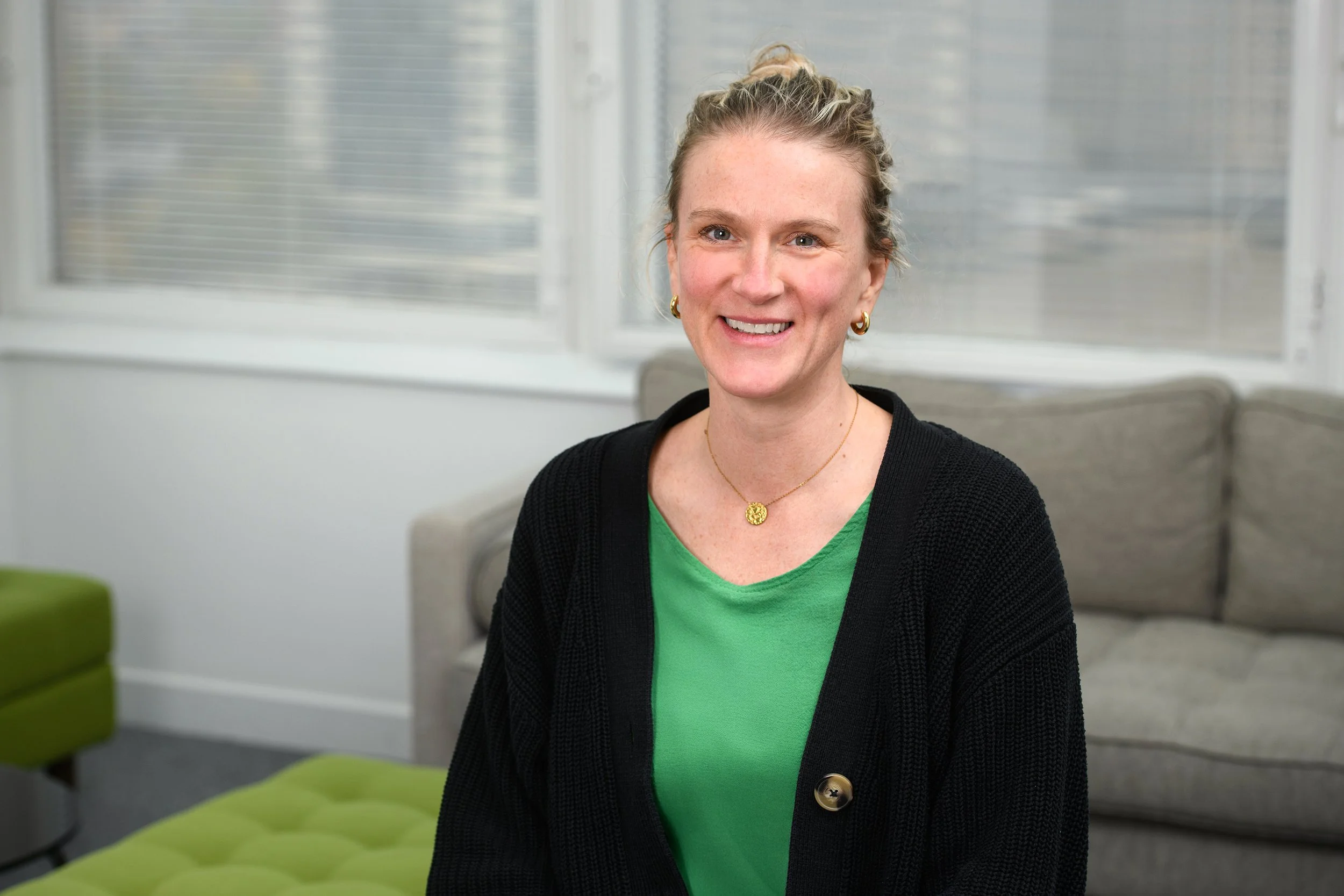 A smiling woman with blonde hair tied back, wearing gold hoop earrings, a gold necklace, a green top, and a black cardigan, sitting in a bright room with large windows and green chairs.