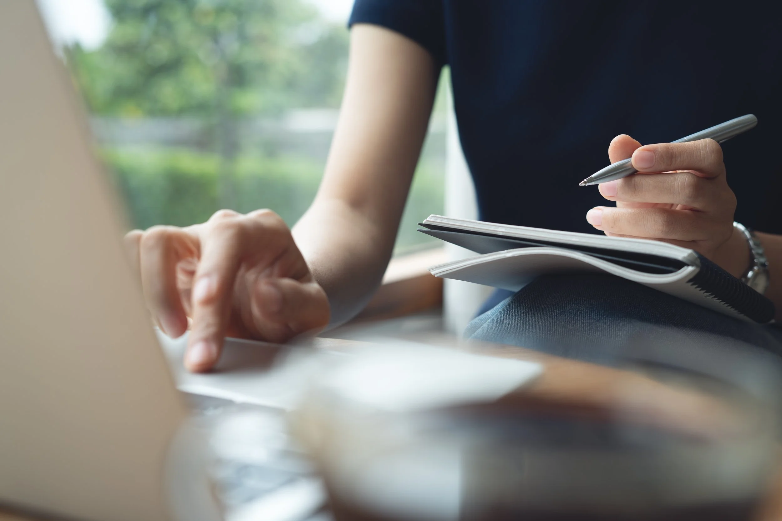 Person holding a pen and writing in a notebook while working on a laptop.