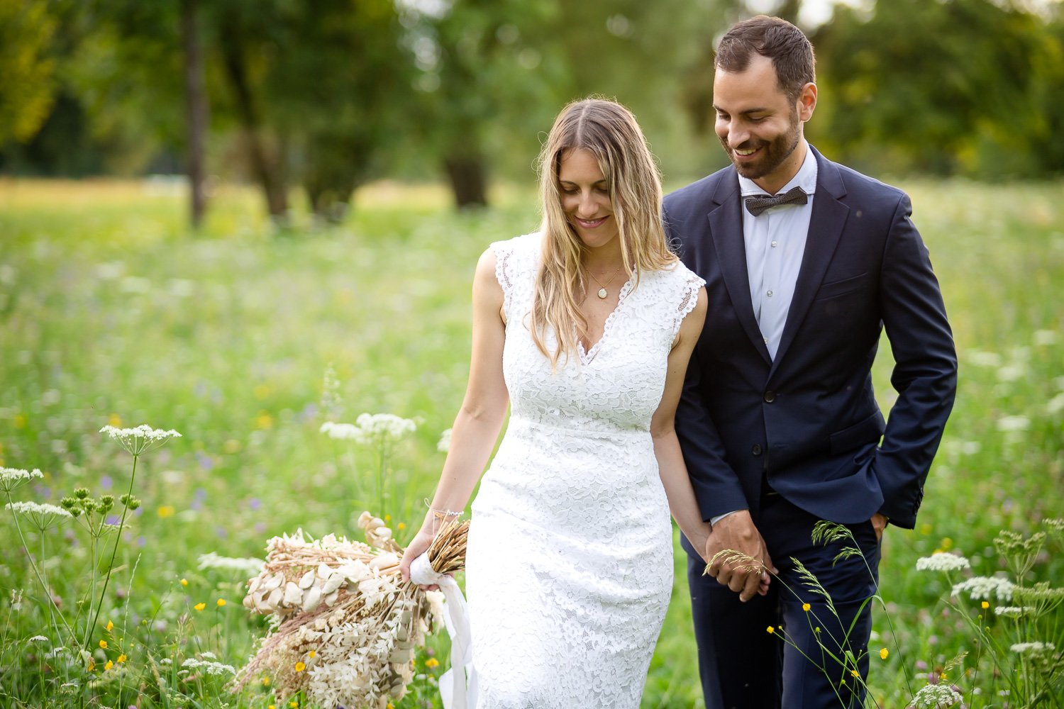 Ein Brautpaar in einem Feld, sie in einem weißen Hochzeitskleid mit Blumenstrauß, er in einem dunklen Anzug. Beide lächeln und halten Händchen. Grünen Hintergrund mit Bäumen und Wildblumen.