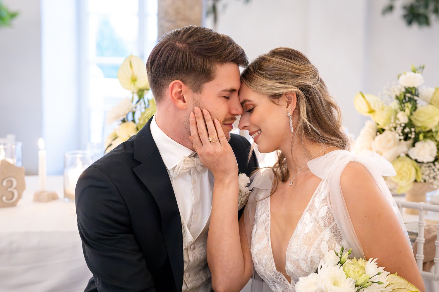 Ein Brautpaar bei ihrer Hochzeit, das sich liebevoll anlächelt, während sie die Köpfe aneinander lehnen. Der Bräutigam trägt einen schwarzen Anzug mit Fliege, die Braut ein weißes Brautkleid. Im Hintergrund sind Blumenarrangements und Kerzen zu sehen