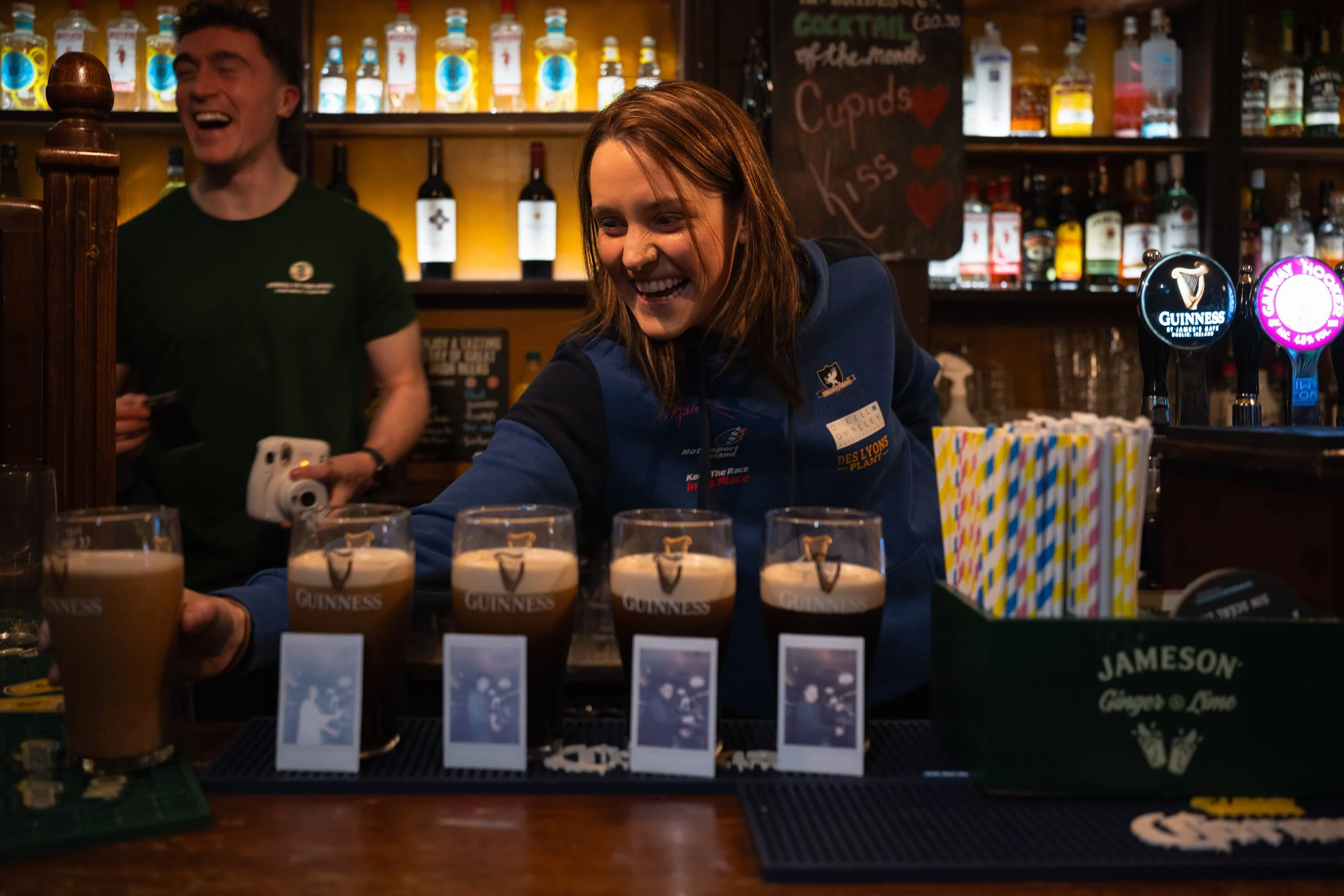 A woman serving multiple pints of Guinness beer at a bar, with a man smiling in the background.