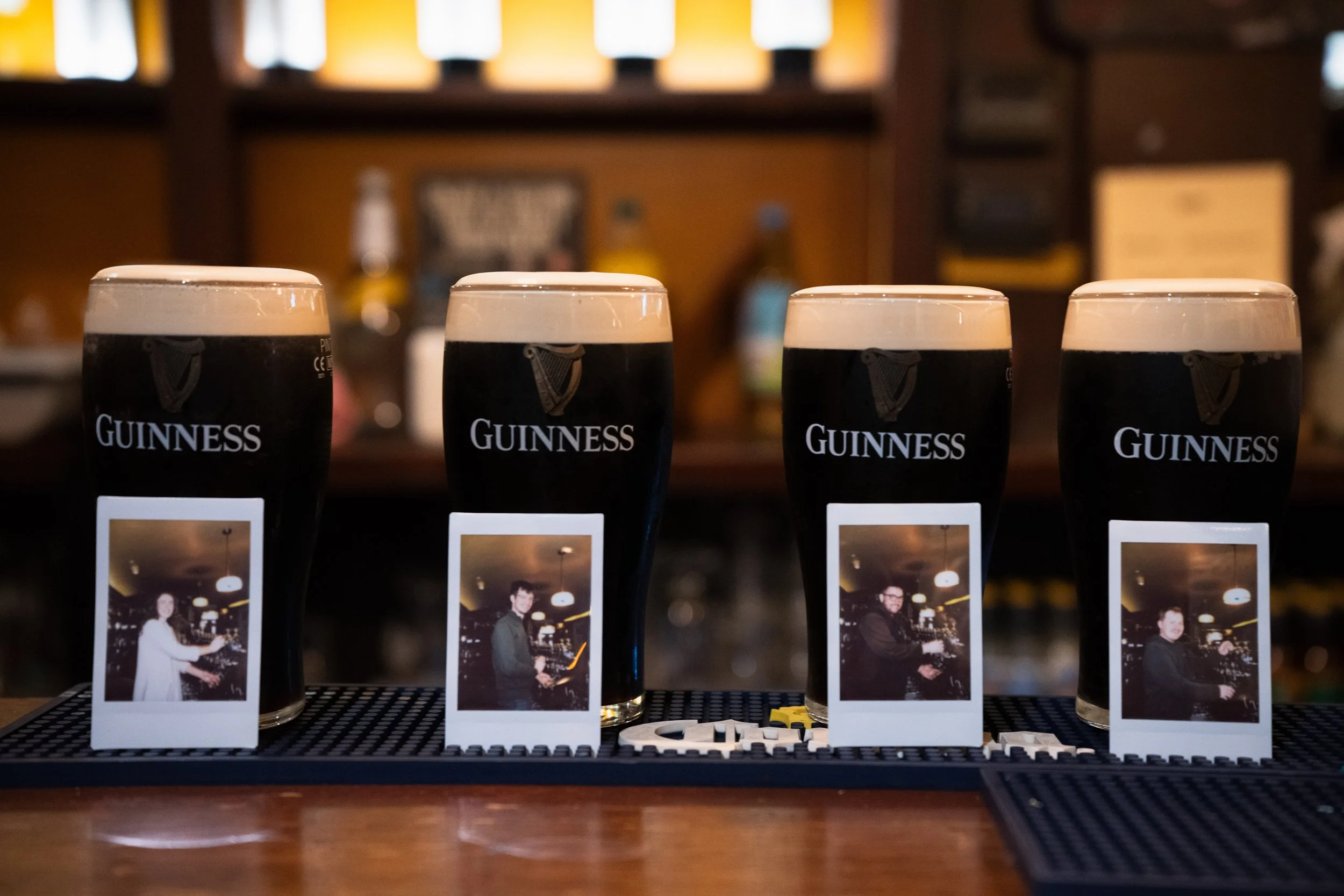 Four Guinness pints with foam on top, each with a Polaroid photo of a different person in front of them, placed on a bar mat.