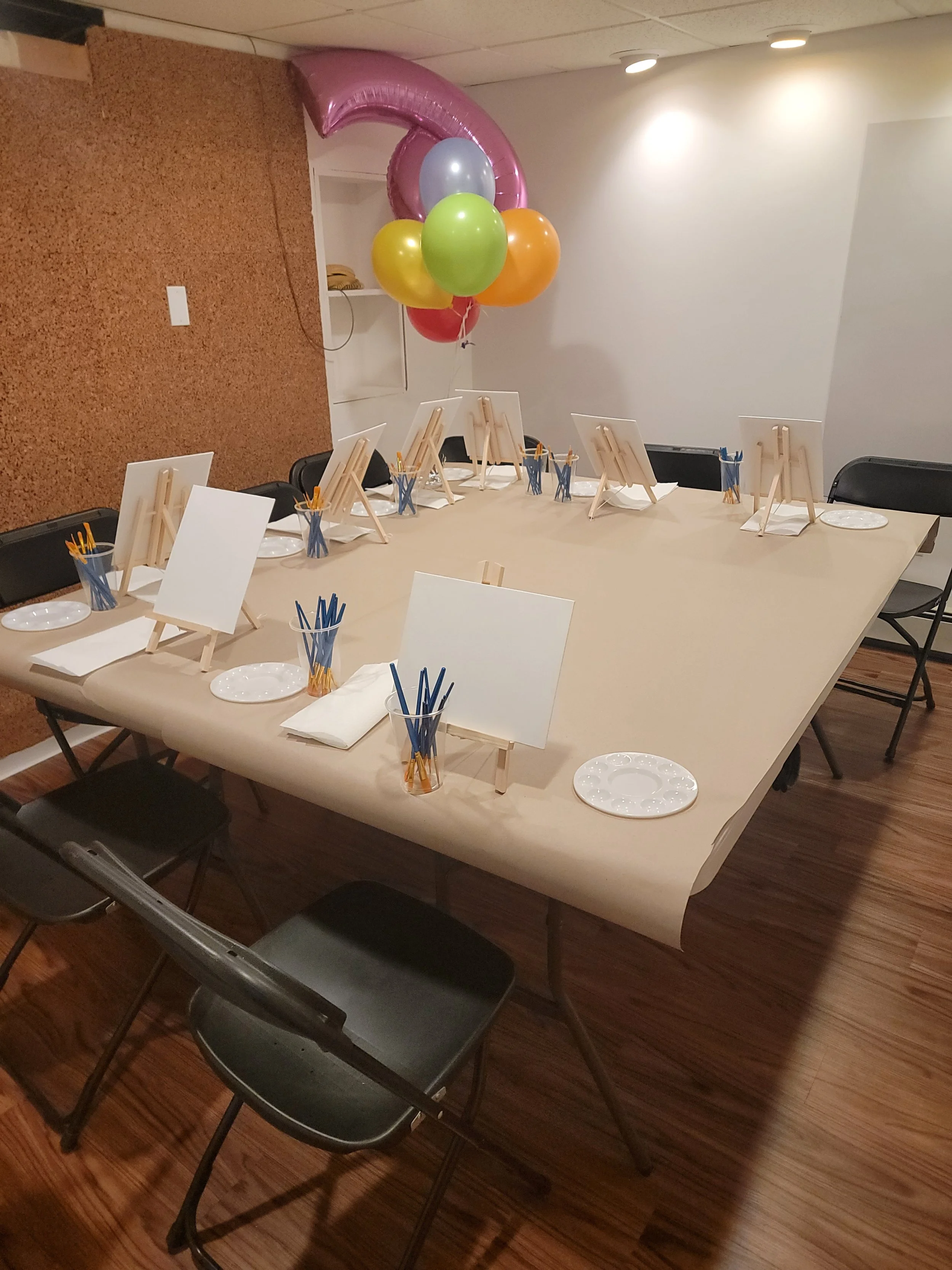 Party room setup with a large table covered with beige paper, small easels, plates, napkins, cups filled with straws, and color balloons, including a pink number '2' balloon, indicating a celebration for a second birthday.