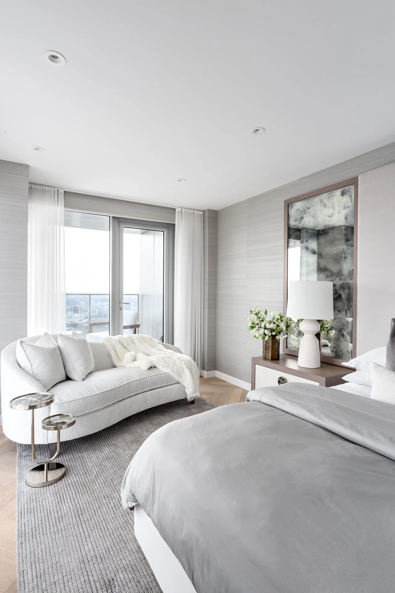 A modern bedroom with a large bed covered in white bedding, a gray sofa with pillows and a faux fur throw, a side table with a metallic lamp, a sliding glass door leading to a balcony, and a vanity with a lamp and flowers.