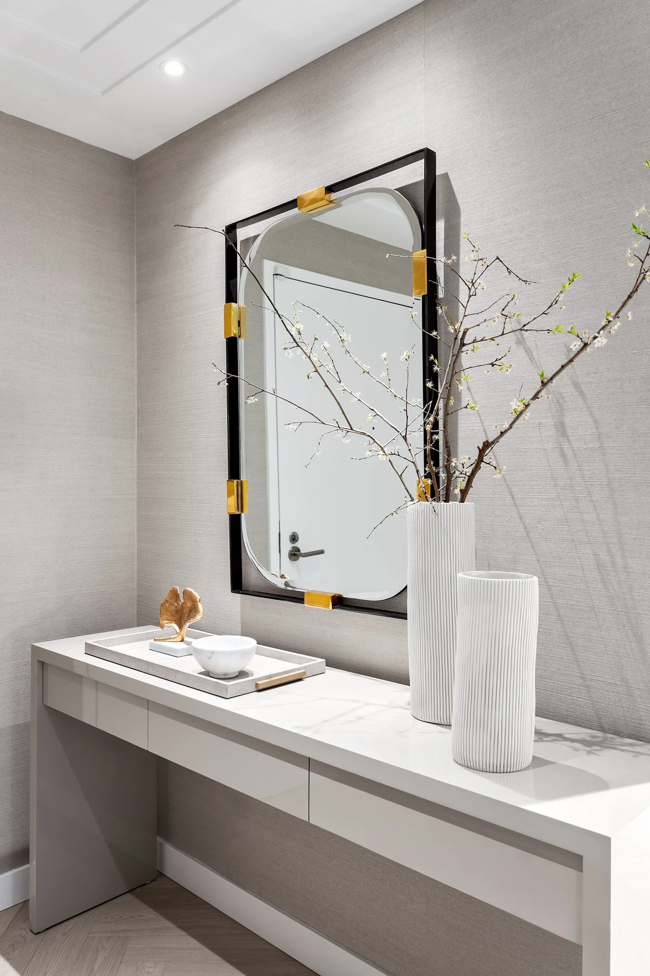 Modern vanity table, a large mirror with a black and gold frame, and decorative vases with branches. The wall is textured gray, and ceiling recessed lighting is visible.