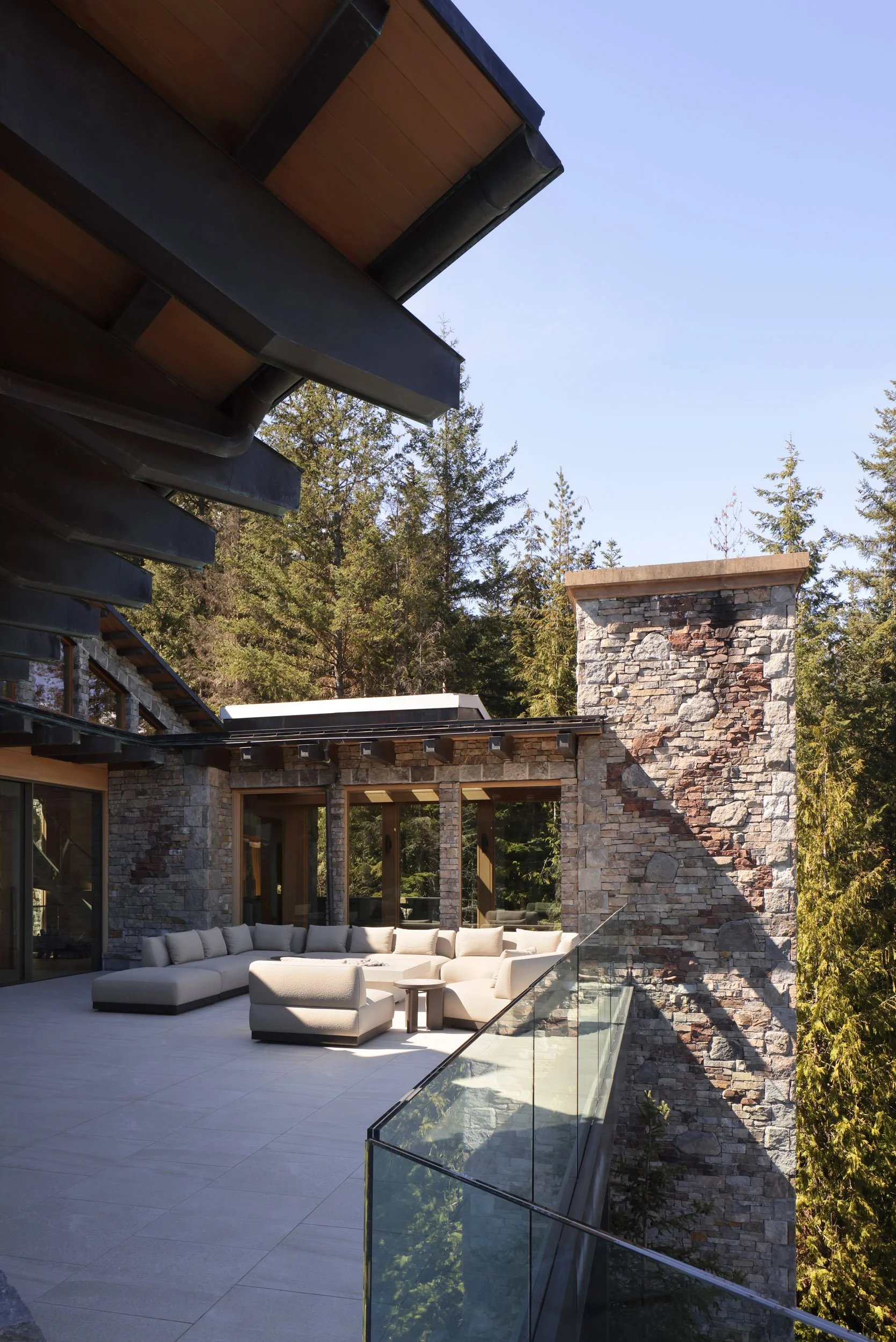 Outdoor patio with modern beige sectional sofa, glass railing, stone walls, and forest background.