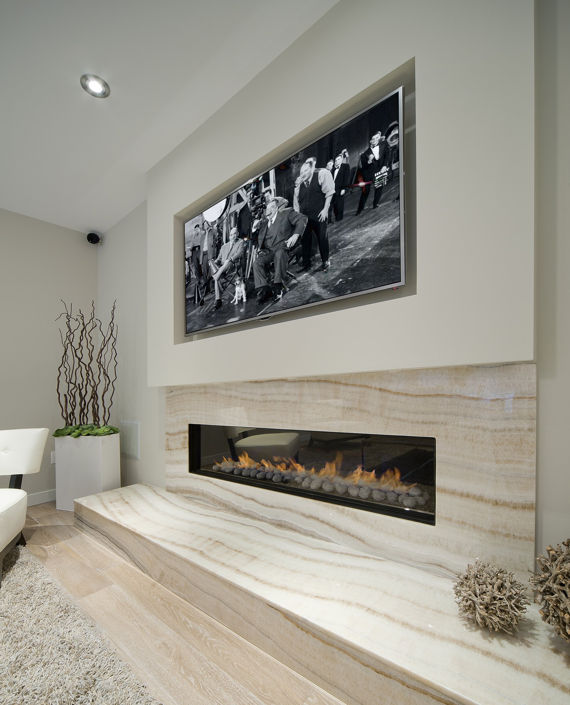 Modern living room with a wall-mounted TV showing a black-and-white photo of men in suits, a gas fireplace below the TV, and decorative plants and objects in the corner.
