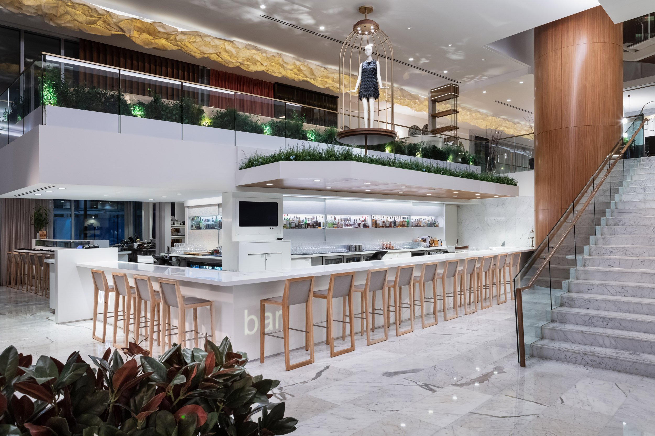 Modern bar with marble flooring, a white counter with high wooden stools, a staircase to an upper level with a mannequin in a cage-like display, and a large wooden pillar. The upper level has green plants and additional seating.