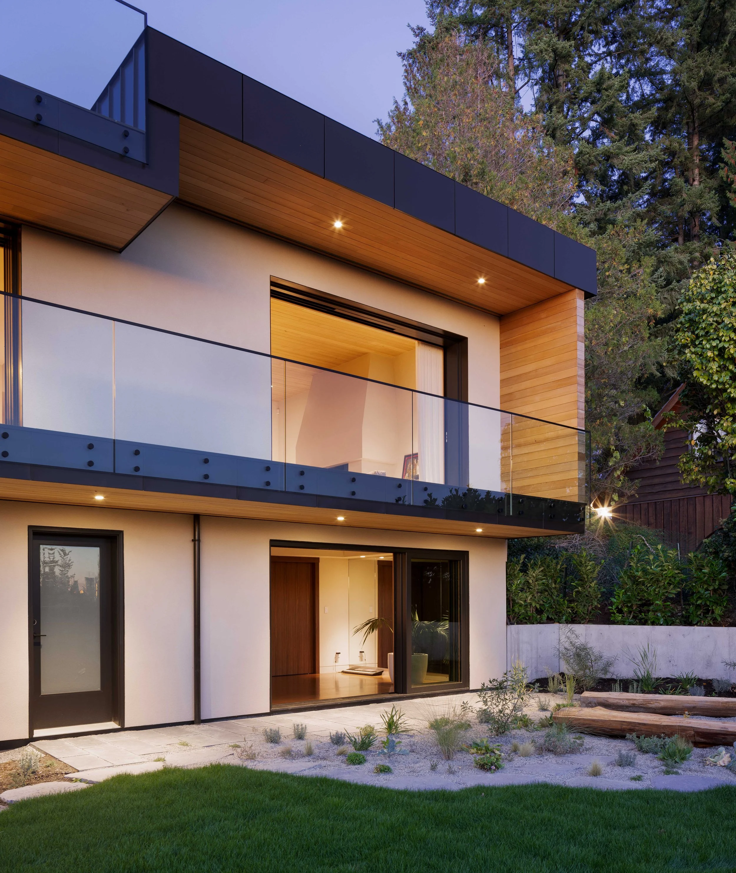 Modern two-story house with large glass sliding doors on the ground floor, a balcony with glass railing on the upper floor, and a landscaped yard with grass, small plants, and wooden decor.