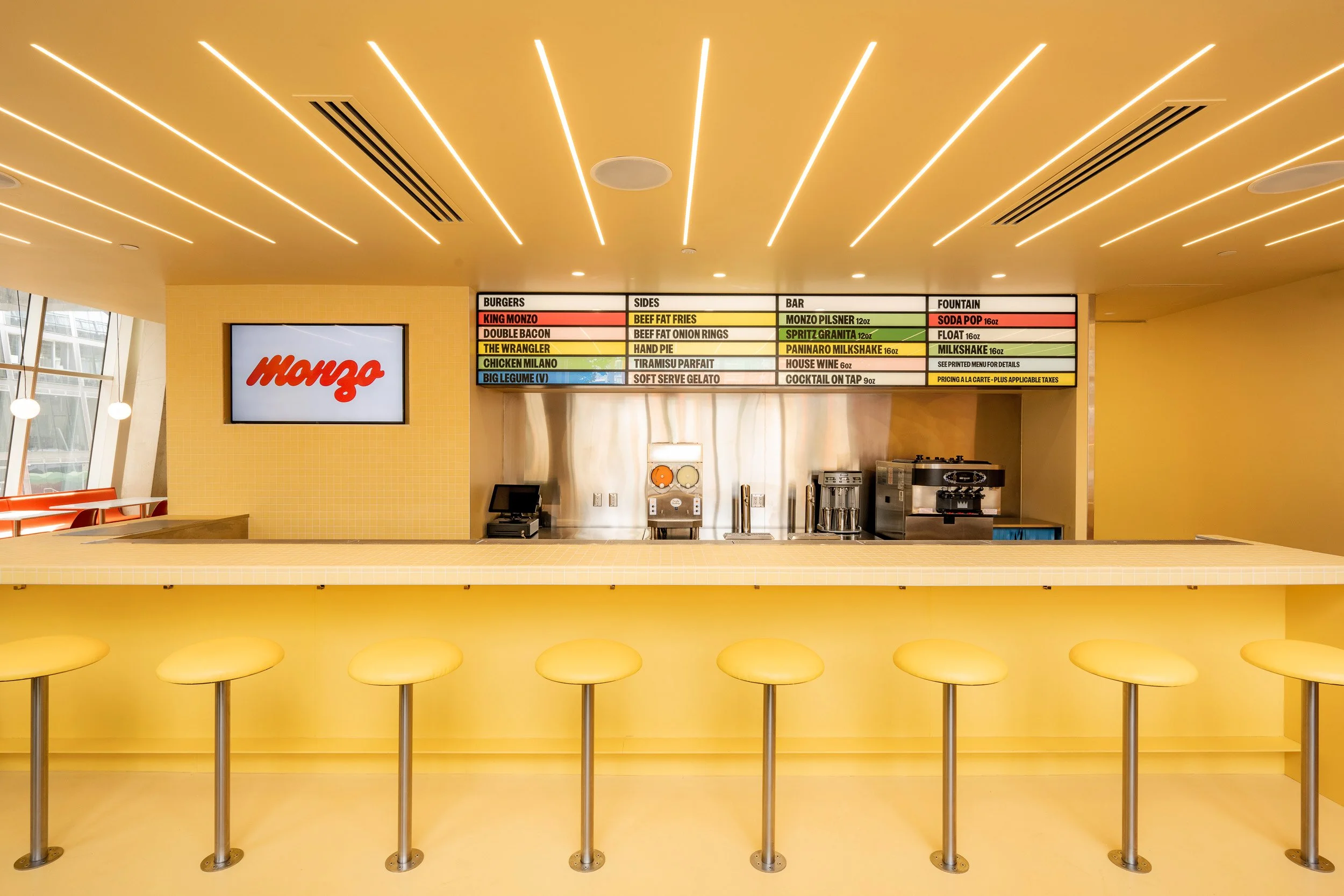 Inside a modern fast-casual restaurant with a bright yellow and yellow color scheme, a counter with five yellow stools, a digital menu board displaying food items, and a large screen with the restaurant's logo, 'Mongo'.