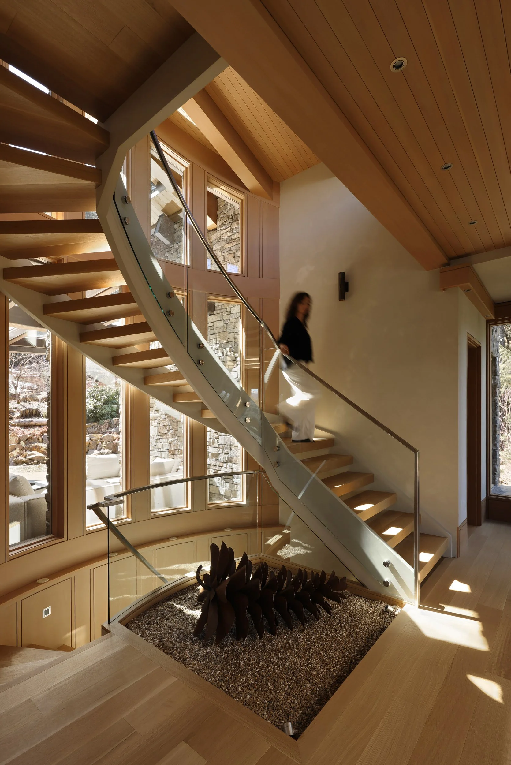 Interior view of a modern home with a curved staircase made of wood and glass, large windows letting in natural light, and a decorative sculpture at the bottom of the stairwell.