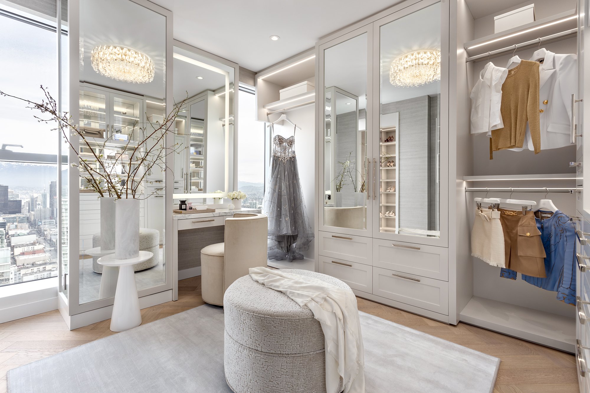 Elegant walk-in closet with white cabinetry, clothing, and accessories, large window with city view, and a dressing table with a mirror.