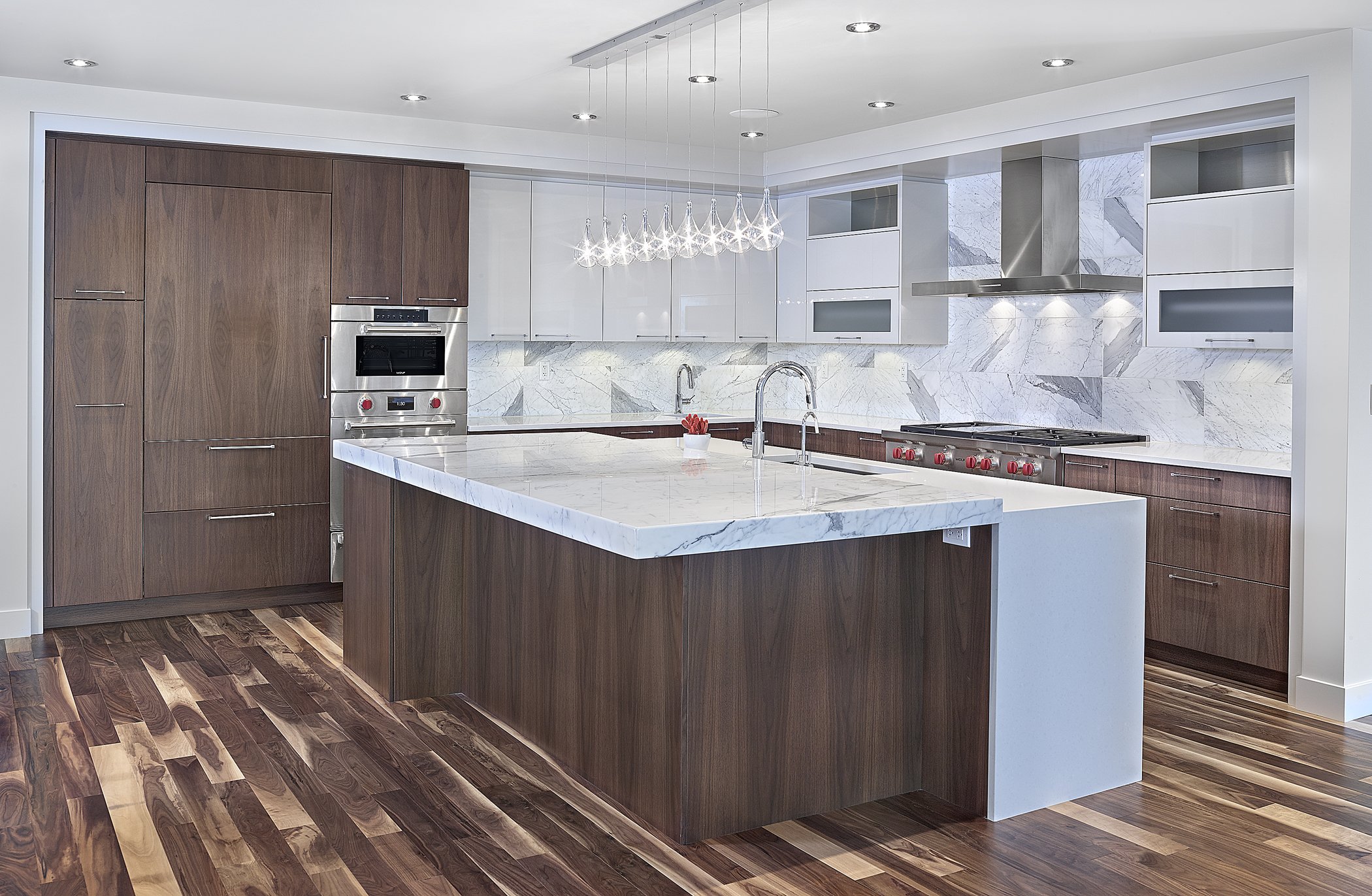 Modern kitchen with wooden cabinets, white marble countertops, and a large kitchen island. Features built-in oven, stainless steel appliances, marble backsplash, and hardwood floors.