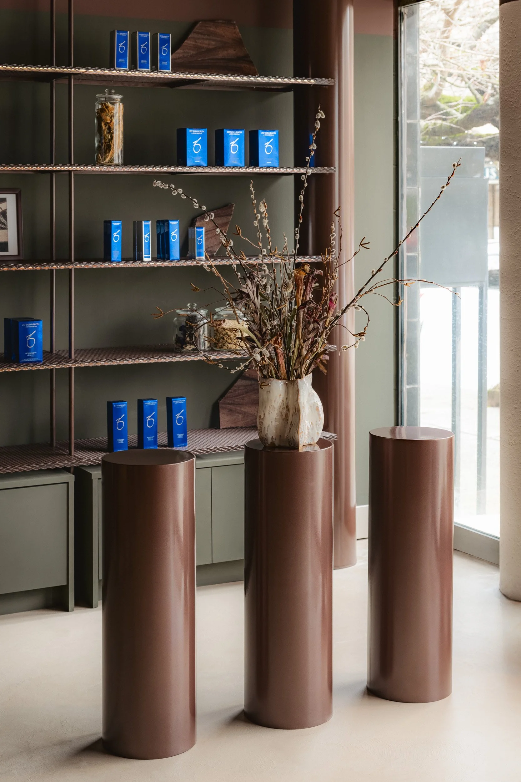 Interior of a modern shop with a green wall, a metal and wood shelving unit holding multiple blue boxed products, a glass jar with dried flowers, and decorative brown cylindrical pedestals with a large vase of dried branch arrangement in the center. 