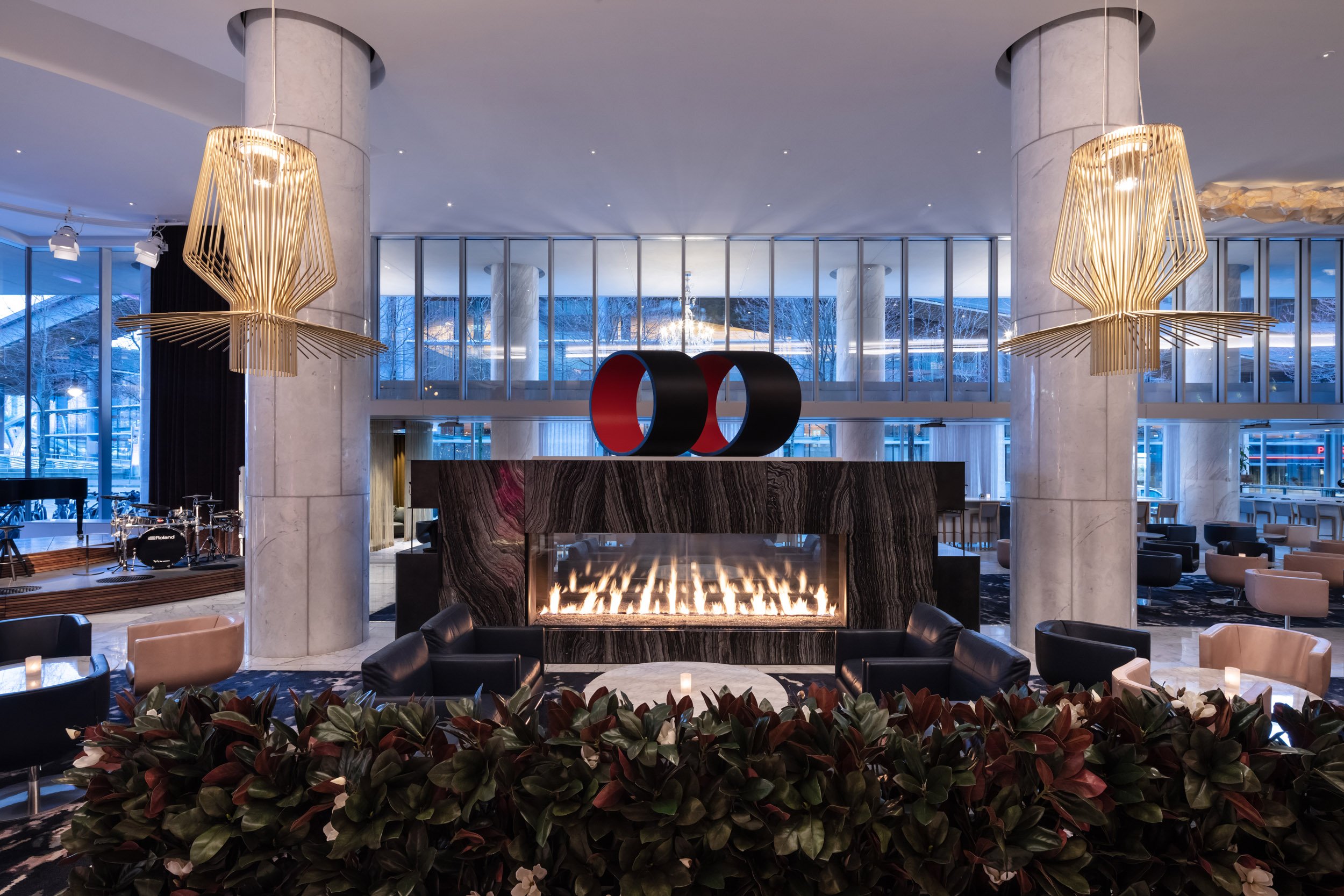A luxurious hotel lobby with a modern fireplace, black and beige armchairs, plants, and large gold chandelier lights. Large windows reveal a city-scape, ocean scene.