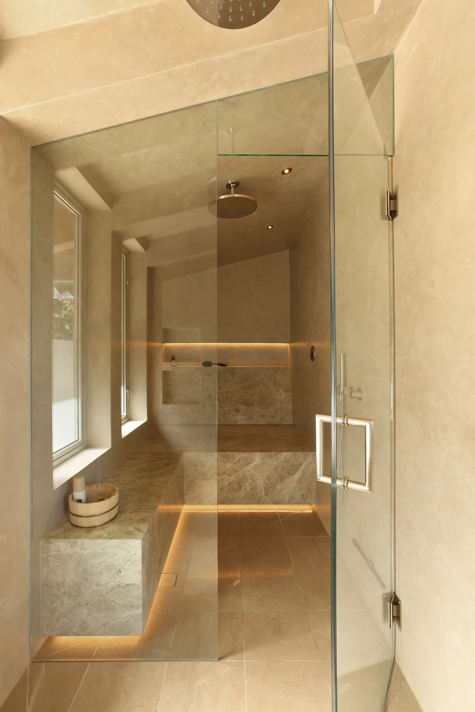 Modern, minimalist shower with beige stone surfaces, glass door, built-in bench, and ambient lighting.