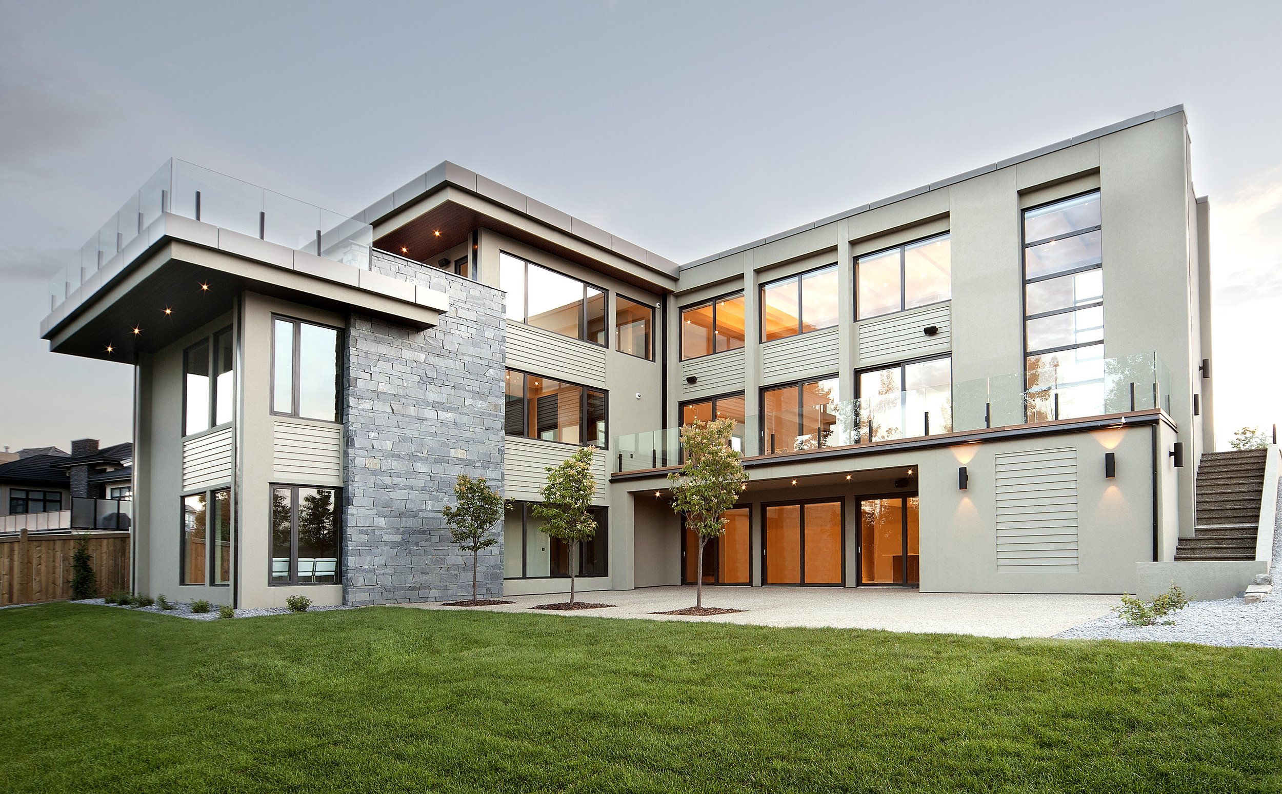 Modern multi-story house with large windows, stone accents, glass balconies, and a well-maintained lawn with small trees.