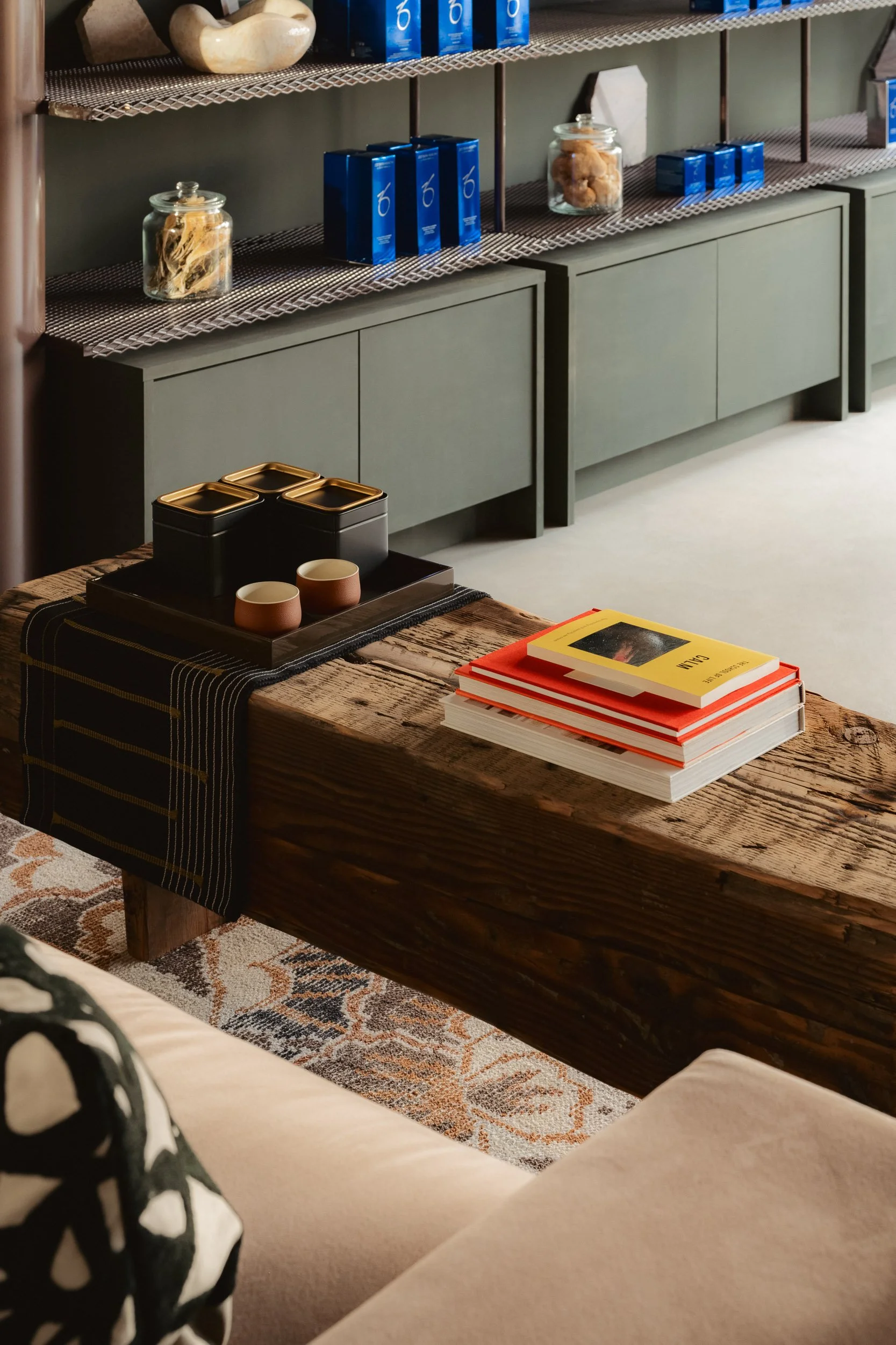 A cozy living room scene with a rustic wooden coffee table, a stack of books, a decorative tray with two small cups, and black containers with gold lids. In the background, a gray sideboard with items on top and metal shelving holding blue boxes, gla
