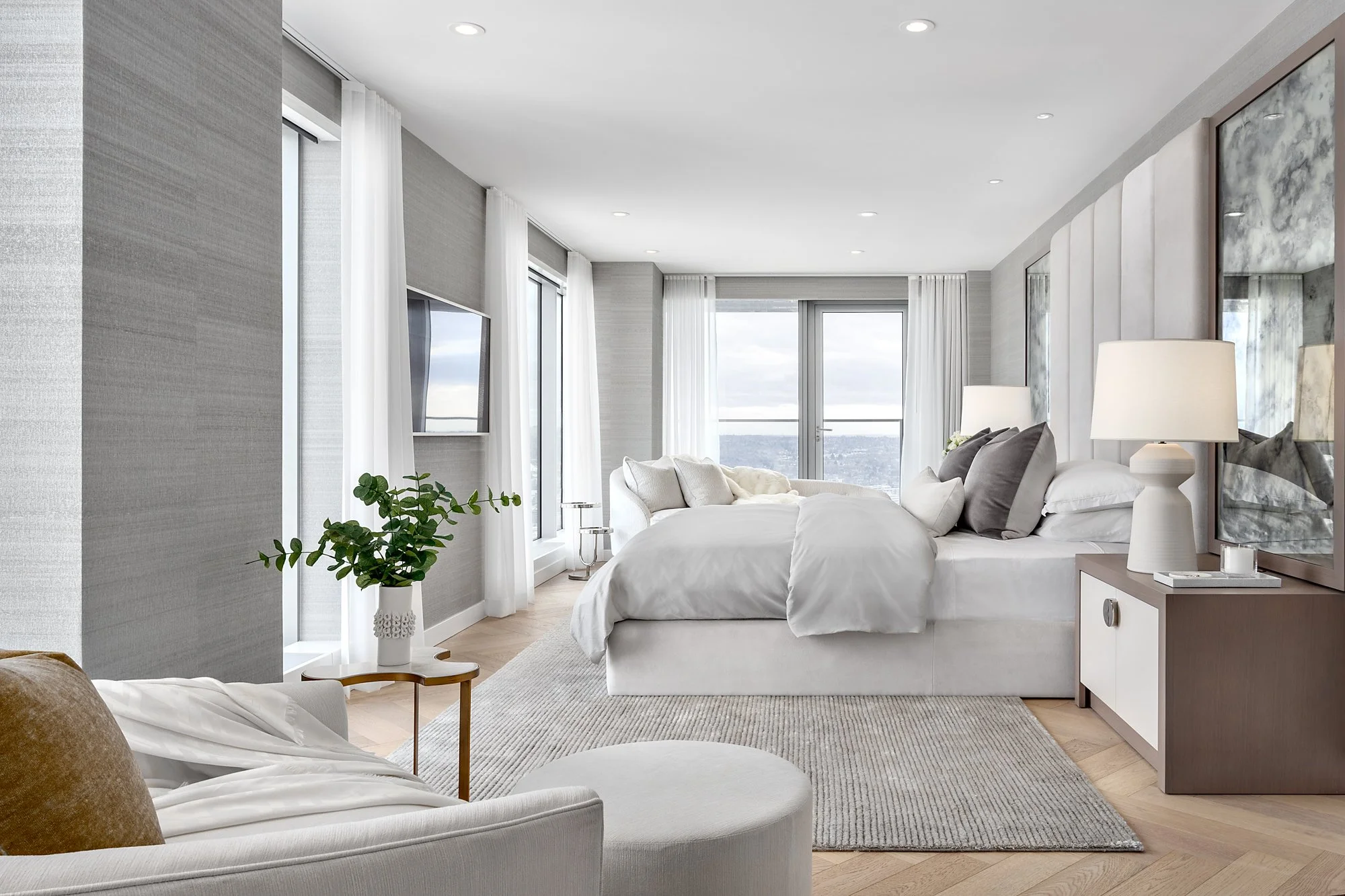Bright, modern bedroom with white bedding, large windows with white curtains, a potted plant, a TV, and a sliding door leading to a balcony with an ocean view.