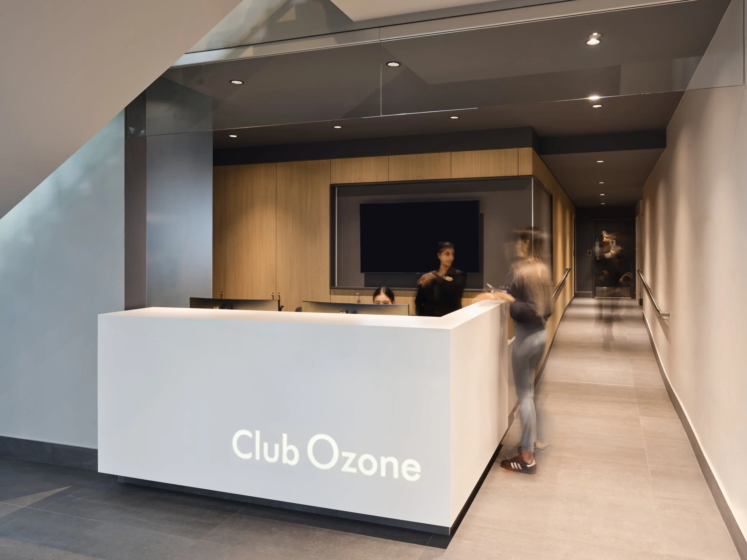 Reception desk at Club Ozone with two staff members behind the counter and a woman standing in front, all inside a modern, sleek lounge area.