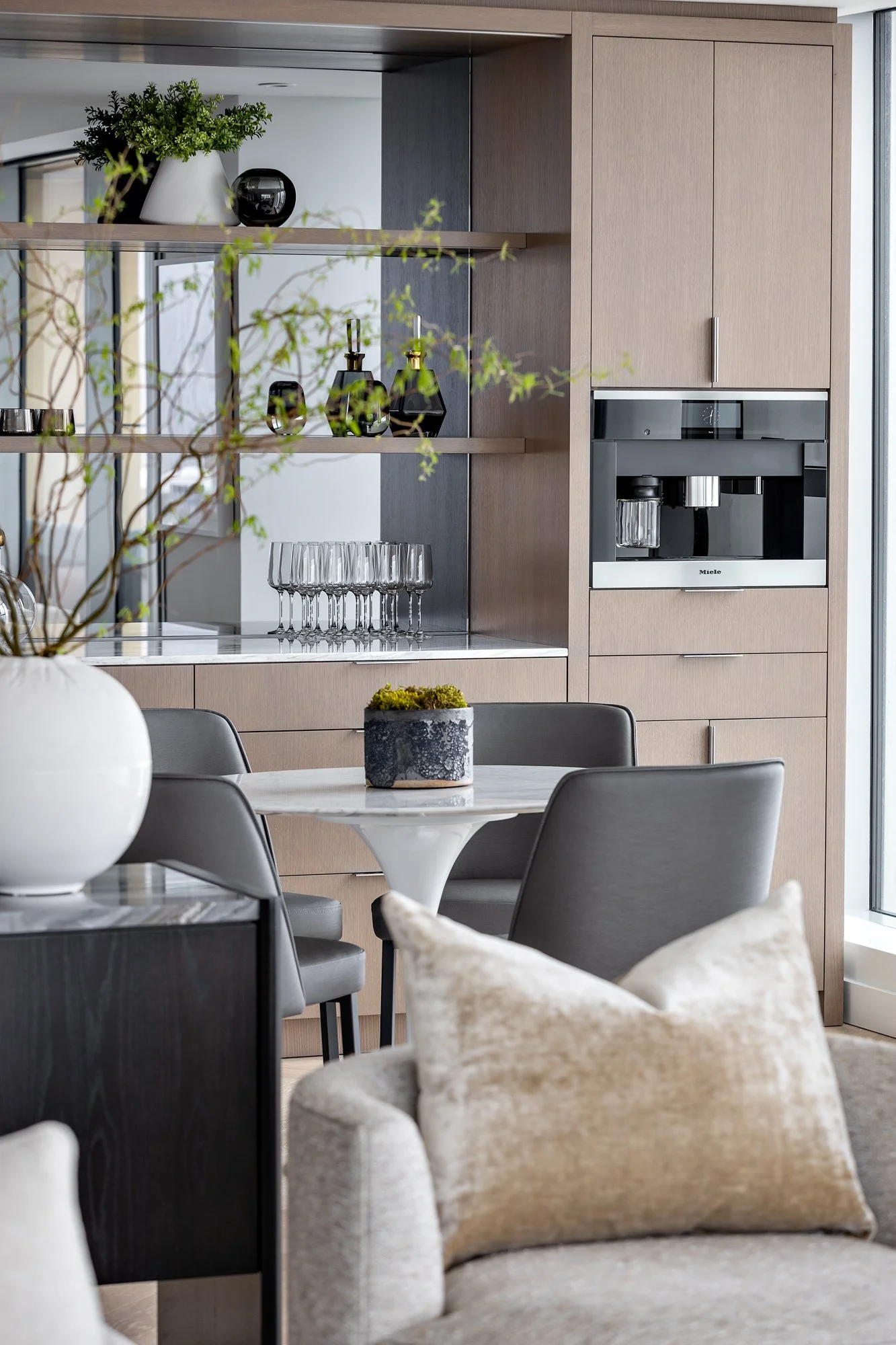 Modern kitchen with light wood cabinets, built-in coffee machine, and a round dining table with grey chairs. Decor includes a large white vase with branches and small dark vases on open shelves, with wine glasses on the counter.