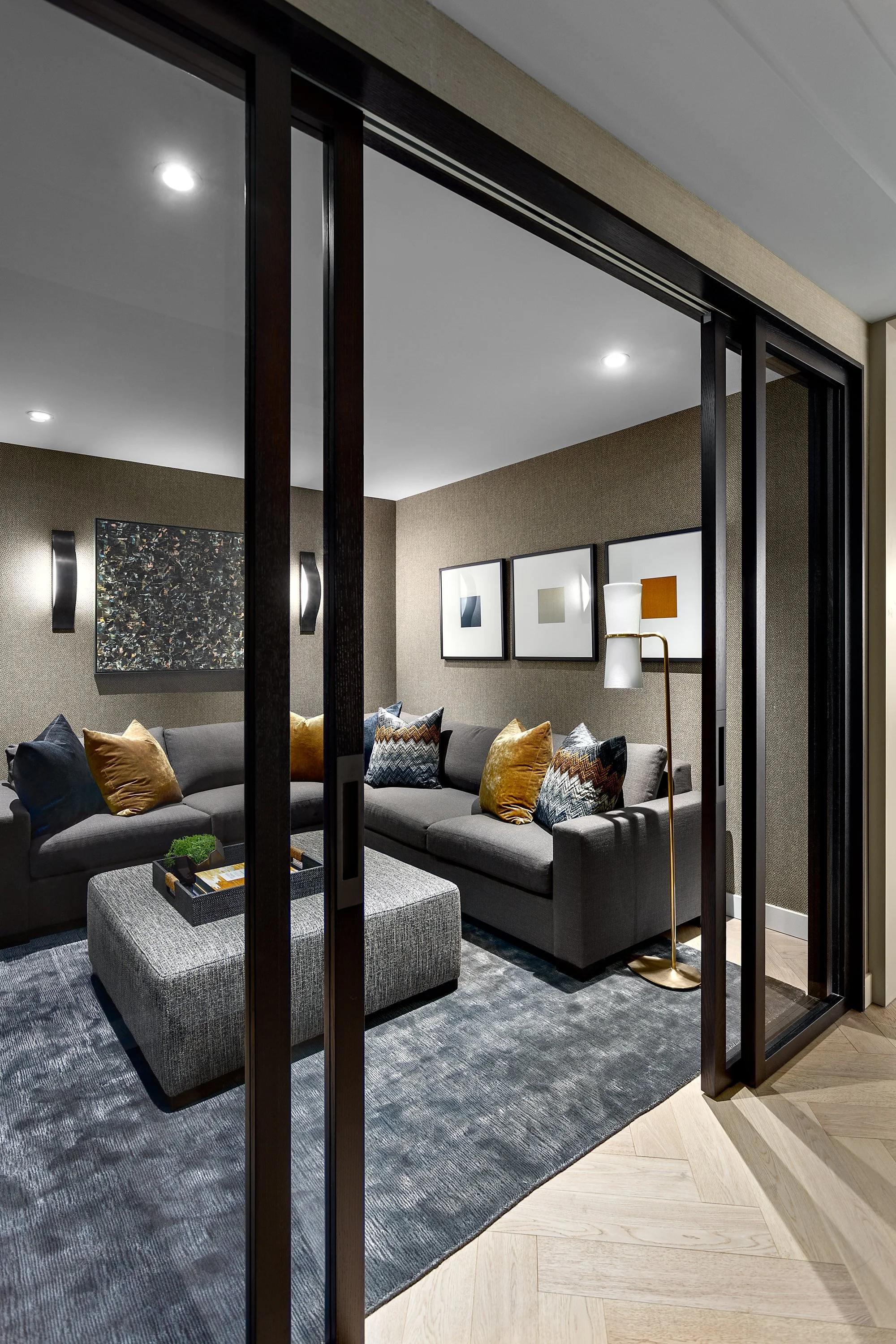 Contemporary living room seen through glass sliding doors, featuring a gray sectional sofa with colorful pillows, framed artwork on the beige textured walls, a gray ottoman with a tray, and a modern floor lamp on a wooden herringbone floor.