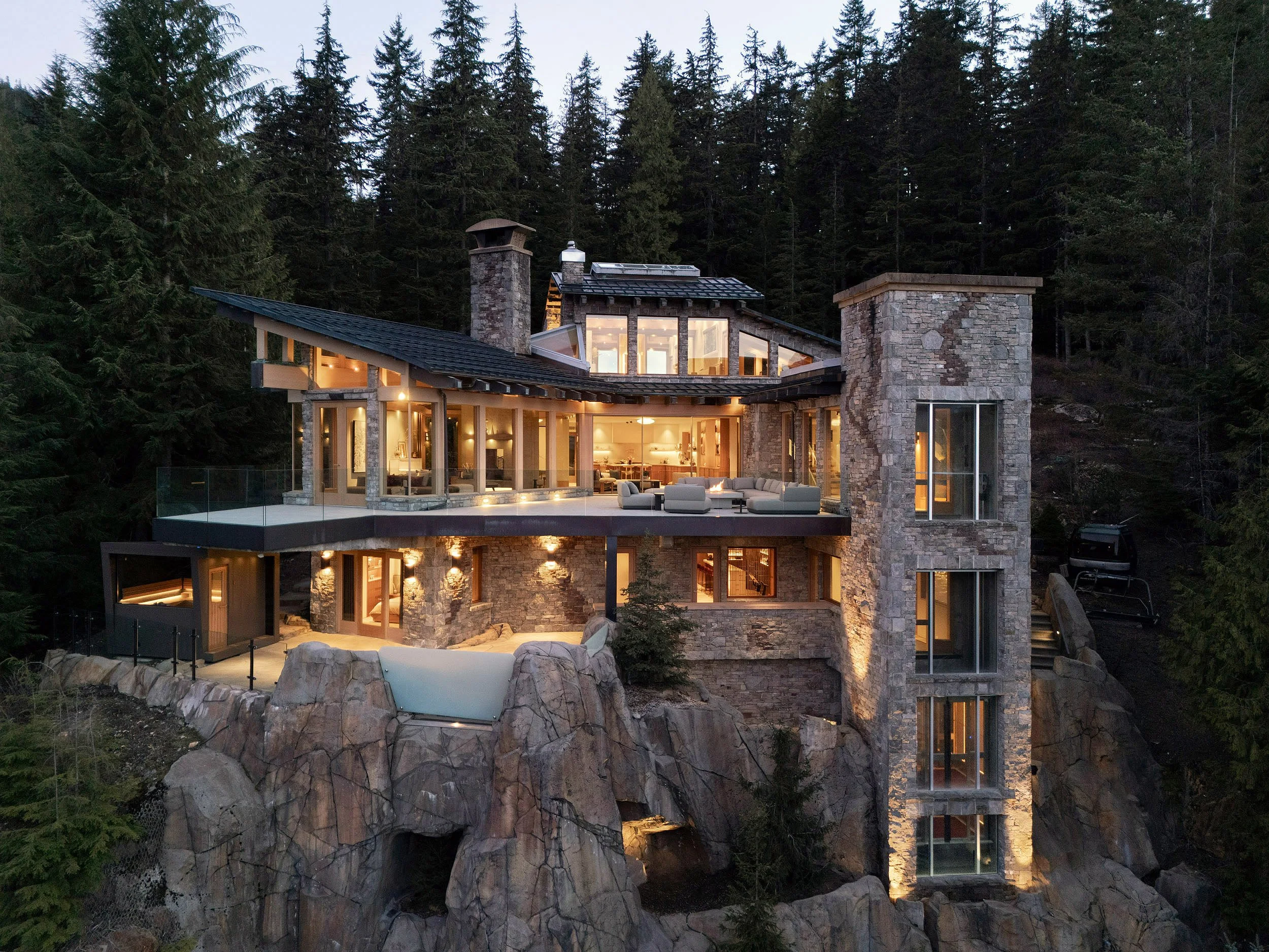 Luxury multi-story house built into rocky hillside with large glass windows and forested background at dusk, illuminated from within.