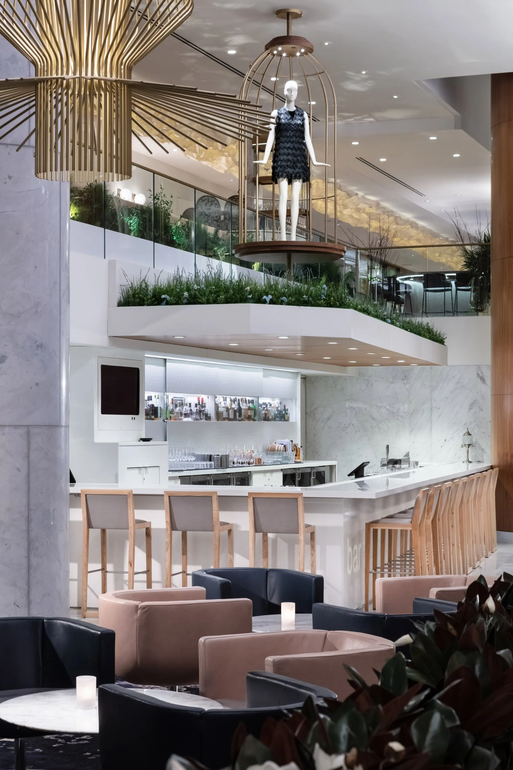 Modern hotel lobby with a bar area, beige and black armchairs, marble tables, and decorative plants. A black mannequin dressed in a black dress is displayed inside a golden birdcage overhead. The space has clean lines, neutral colors, and artistic li