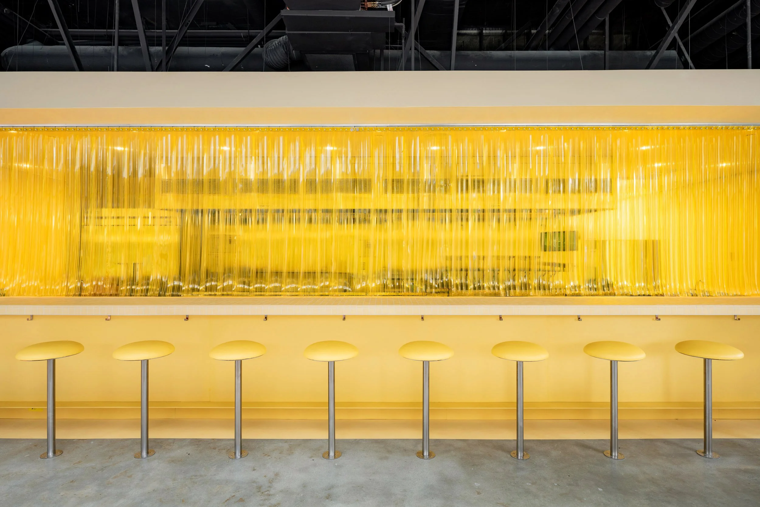 A bright yellow bar counter with yellow bar stools in front and a yellow plastic curtain behind. The setting appears modern and minimalist.