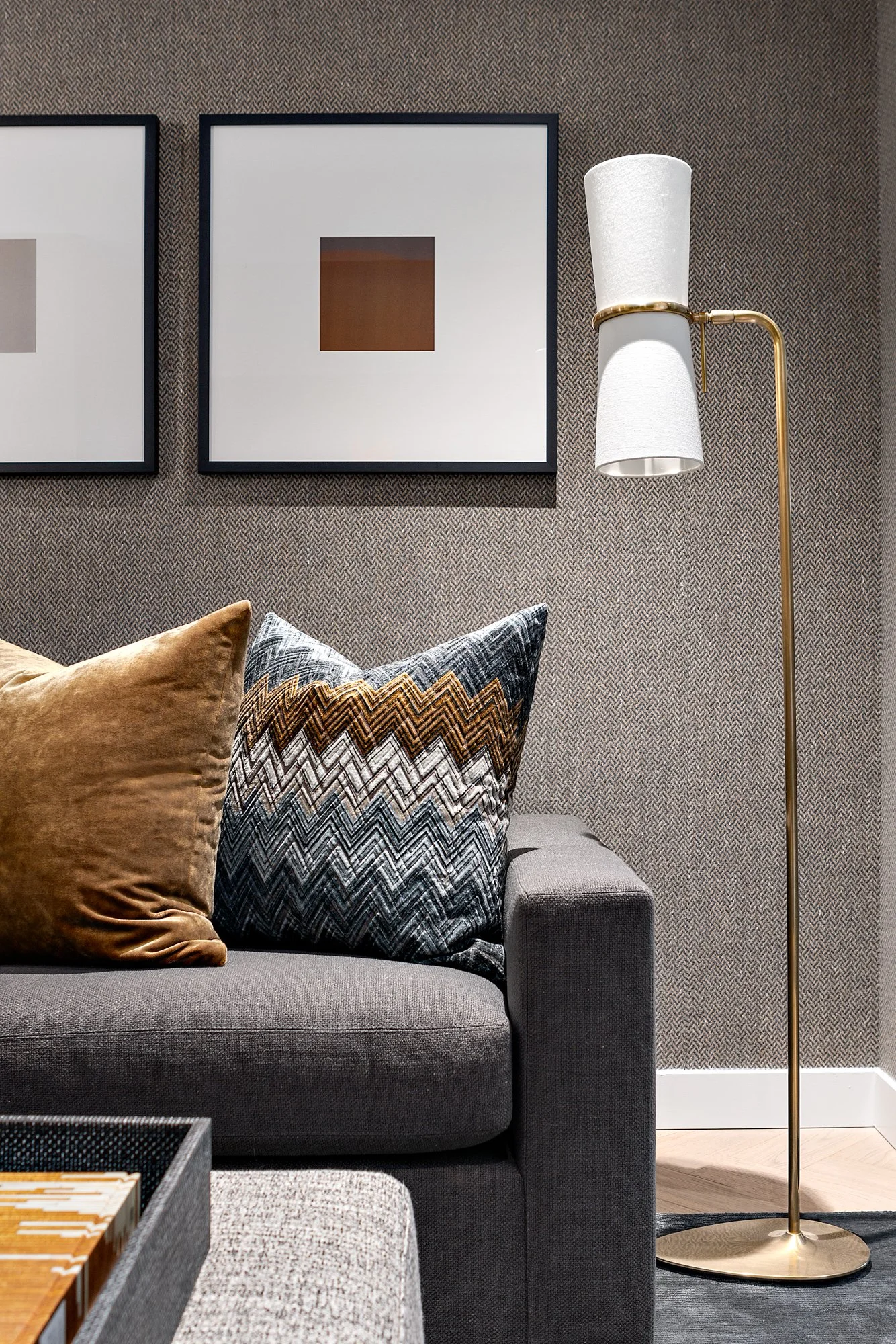 Living room corner with a dark gray upholstered sofa, two decorative pillows (one in mustard and the other with a chevron pattern in black, white, brown, and gray), a textured gray wall with framed abstract art, and a tall white and gold floor lamp w