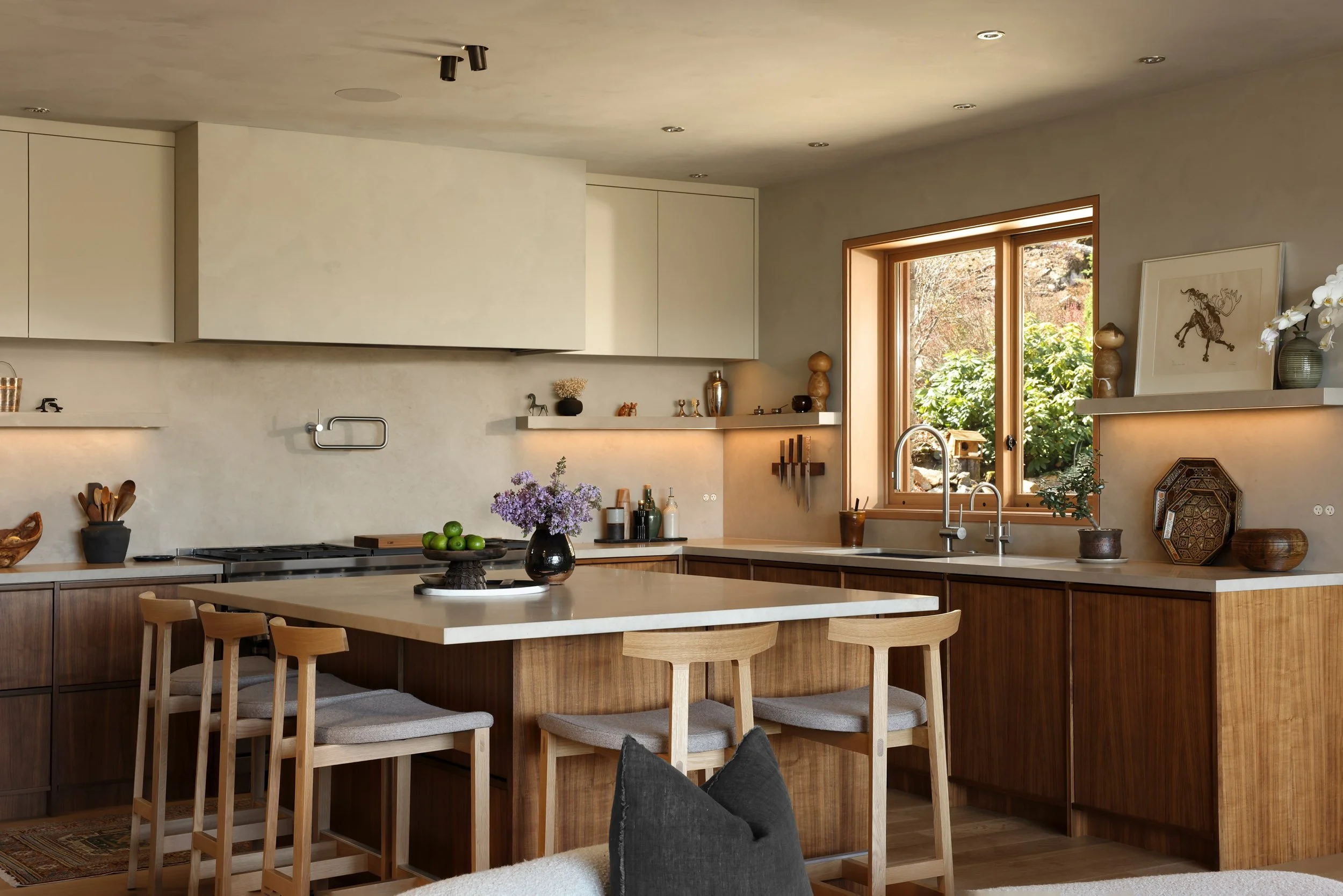 Modern kitchen with wooden cabinets, a large island with a white countertop, and a window showing greenery outside. Decorative items and a vase with flowers are on the shelves and countertop.