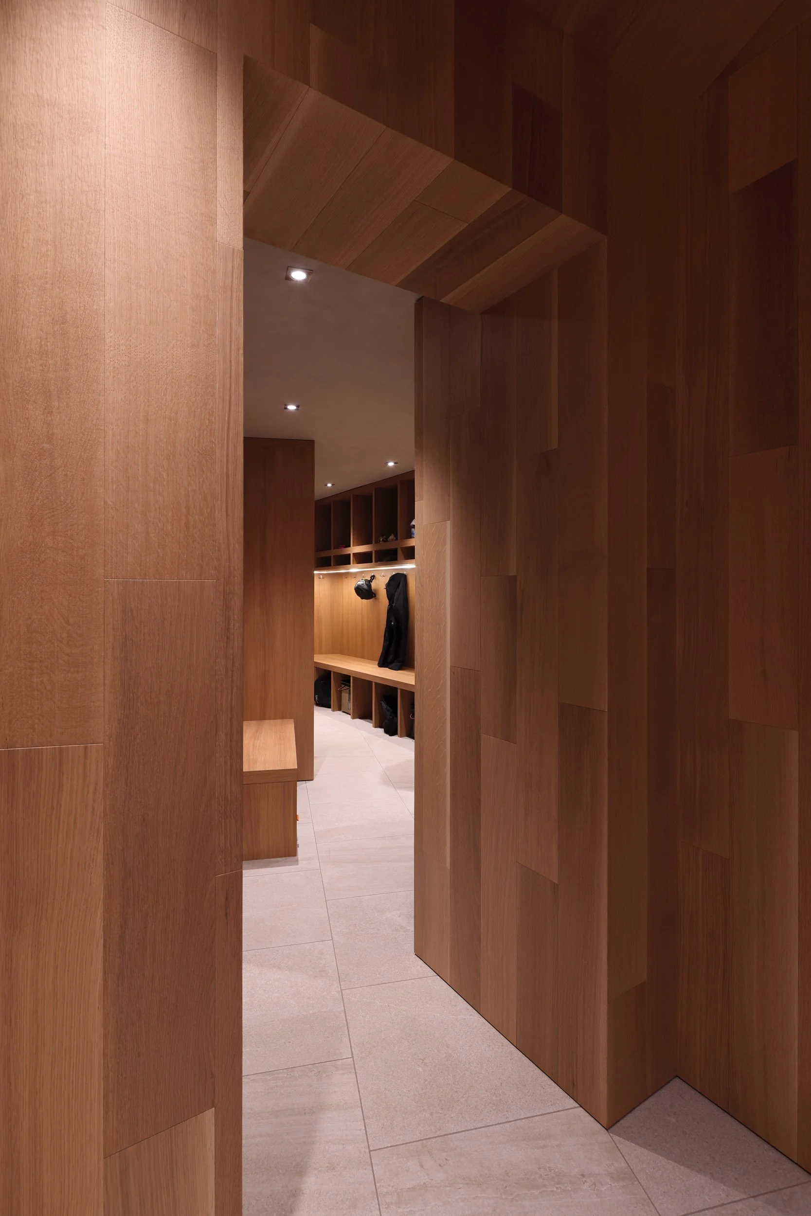 A hallway with wood-paneled walls and built-in storage cubbies, some holding shoes, with a bench and black coat hanging on a hook.