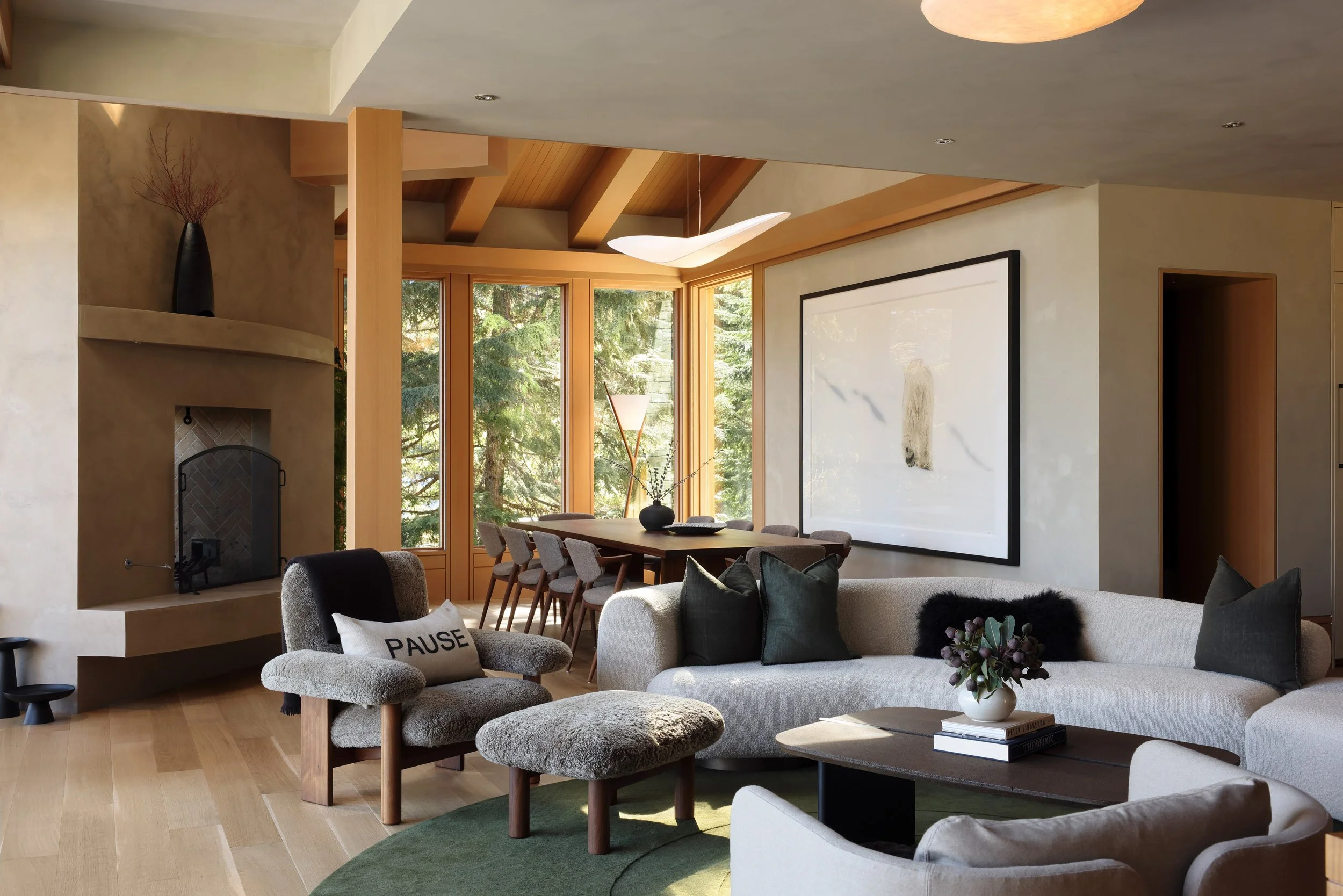 Living room with large beige sectional sofa, black pillows, and a coffee table with a vase of flowers. There is a cozy gray armchair with a "PAUSE" pillow and a matching ottoman. A dining table with chairs is in the background near large windows with