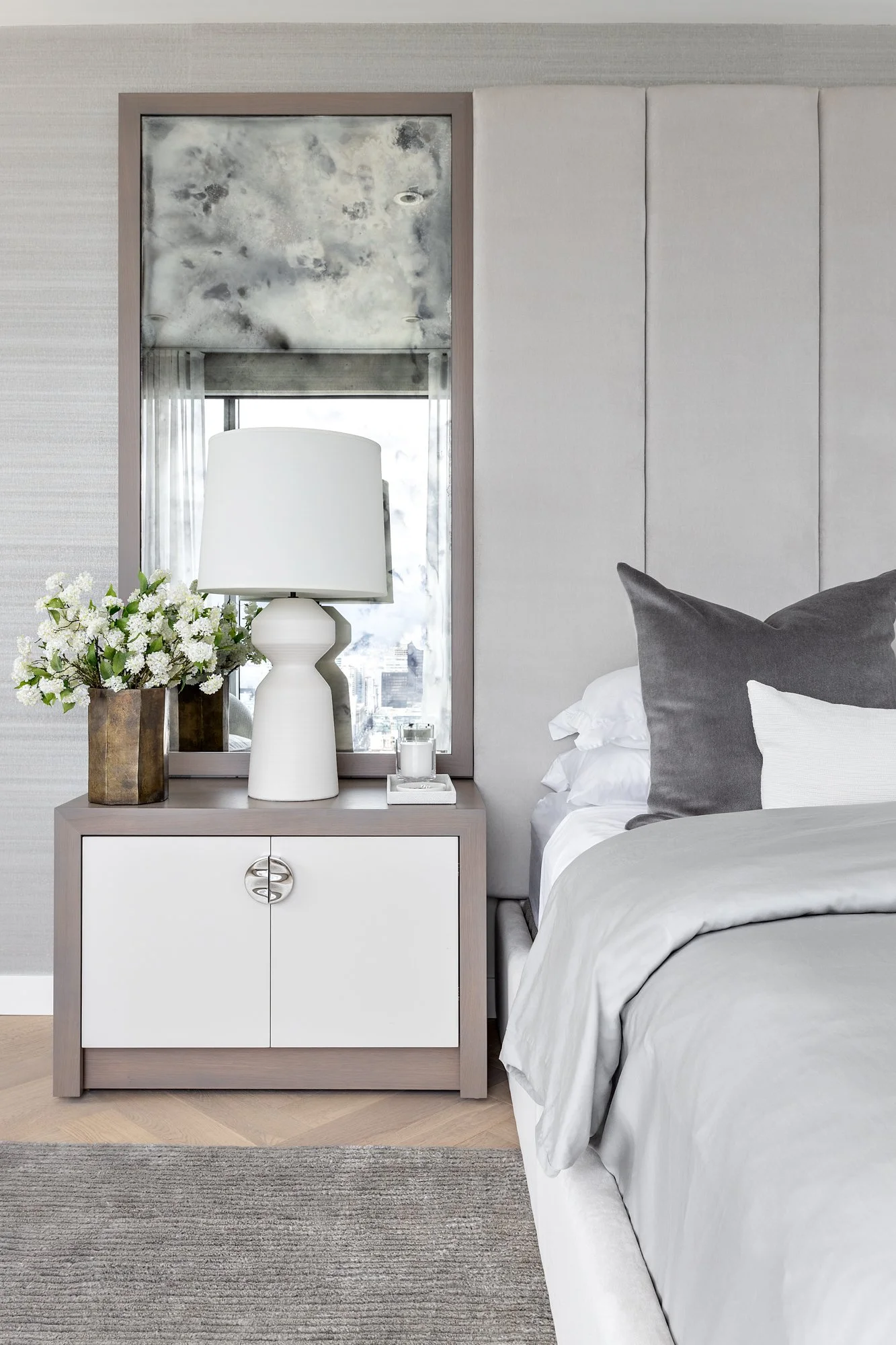 Modern bedroom with a nightstand, white bedside lamp, vase with white flowers, candles, bed with gray and white pillows, and window with city view.