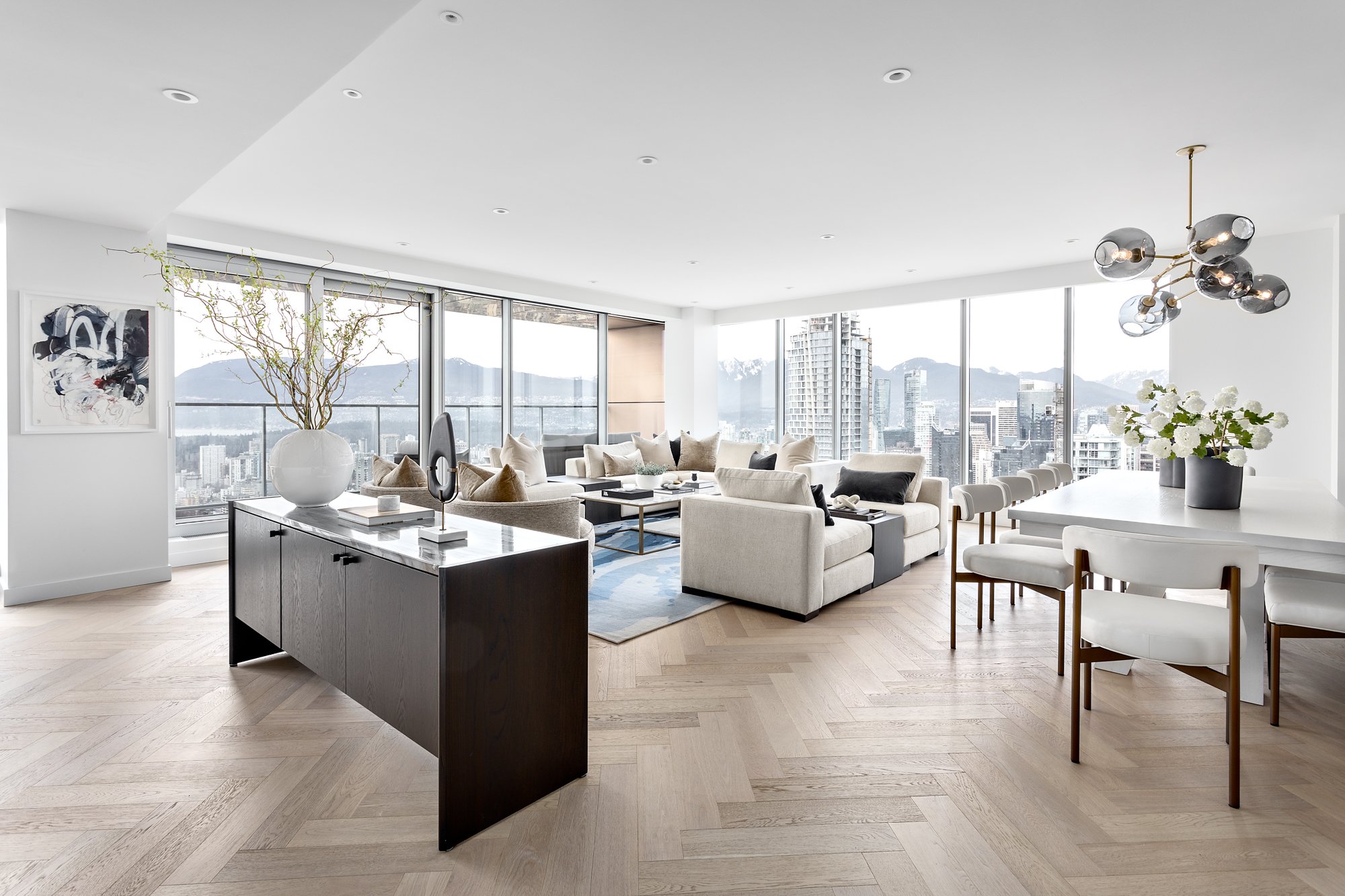 Modern high-rise apartment living room with large windows showing a city skyline and mountains, white sofas, a black sideboard with a large white vase and decorative items, a dining area with white chairs and a table with a flower vase, contemporary 