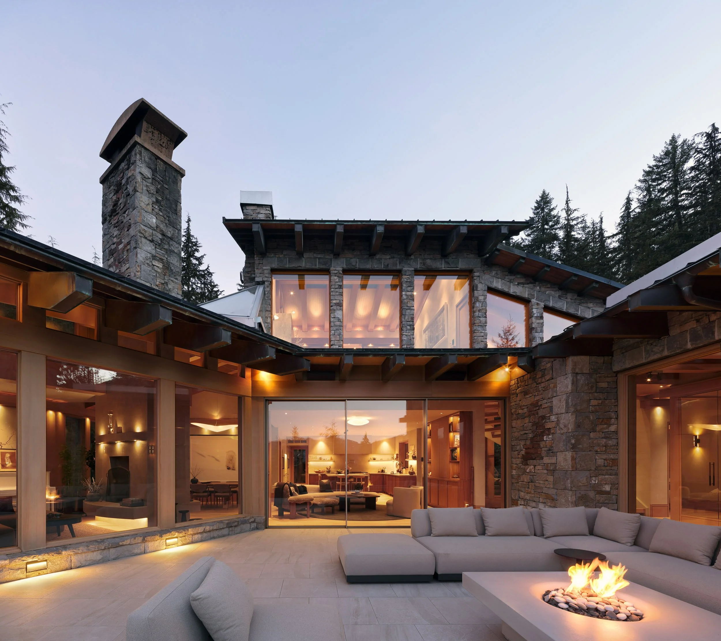 Exterior view of a modern house with large glass windows and a stone facade, showing a cozy outdoor patio with a fire pit, located in a wooded area during dusk.