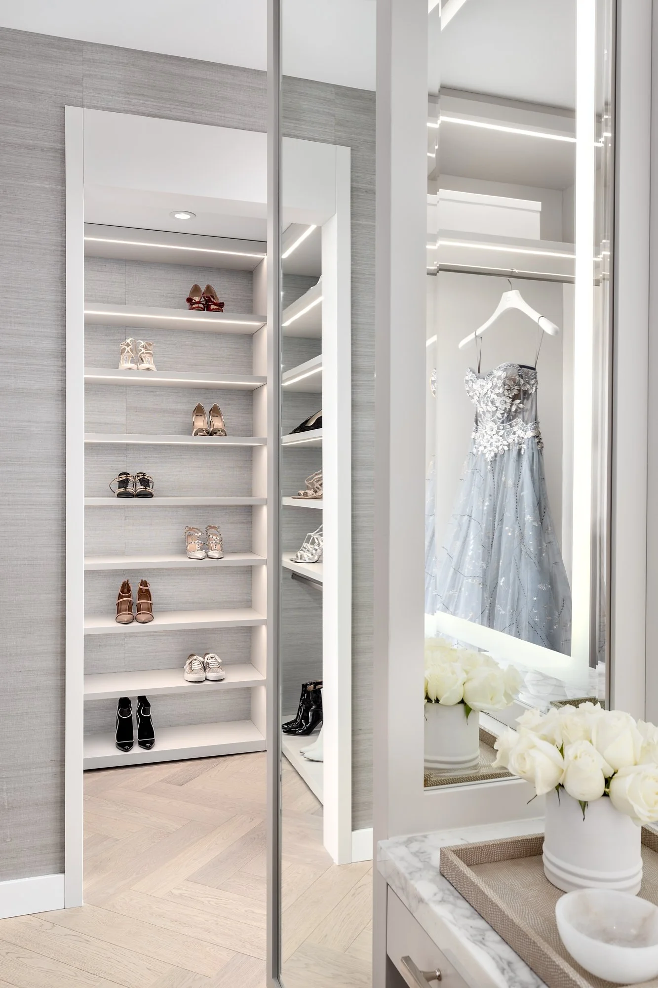 A walk-in closet with shelves of shoes on the left and a display of a dress on the right with white roses on a marble counter in front.