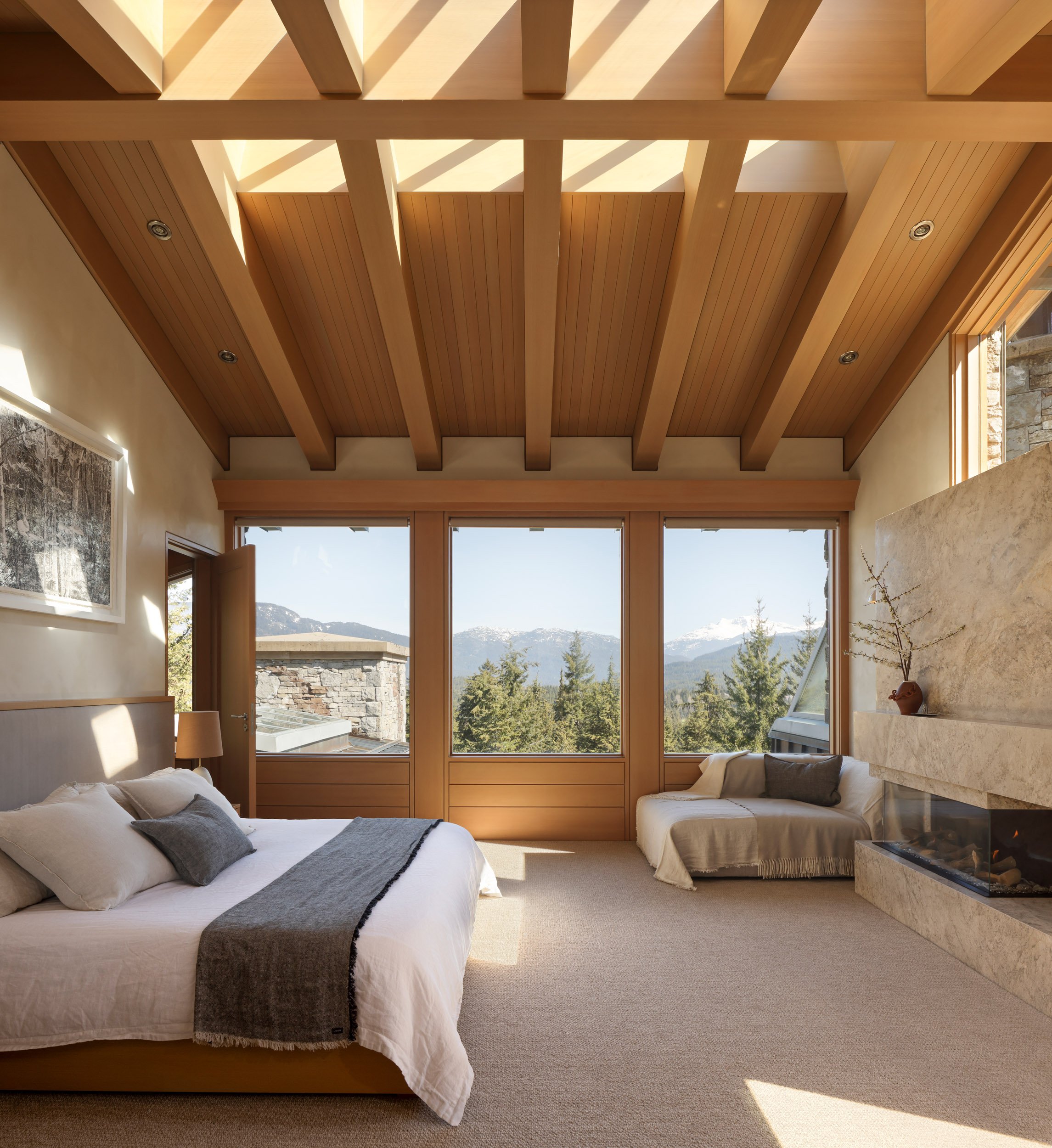 A bedroom with a vaulted wooden ceiling, large windows showing mountain views, a bed with white and gray bedding, a fireplace, and a small seating area.