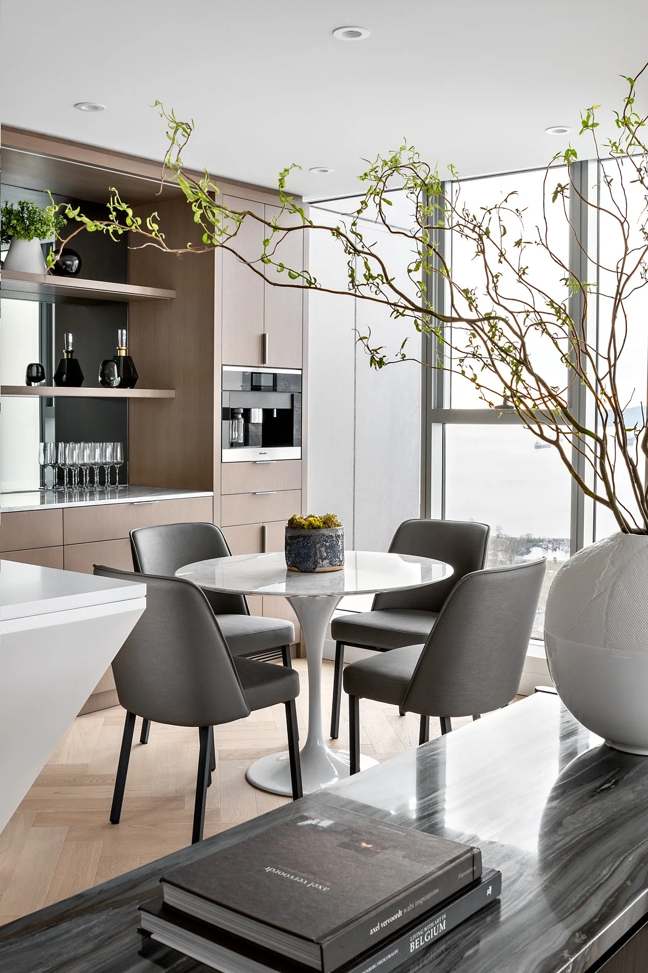 Modern dining area with round white marble table surrounded by five grey upholstered chairs, large decorative branches in a white vase, and a view of water outside large windows.