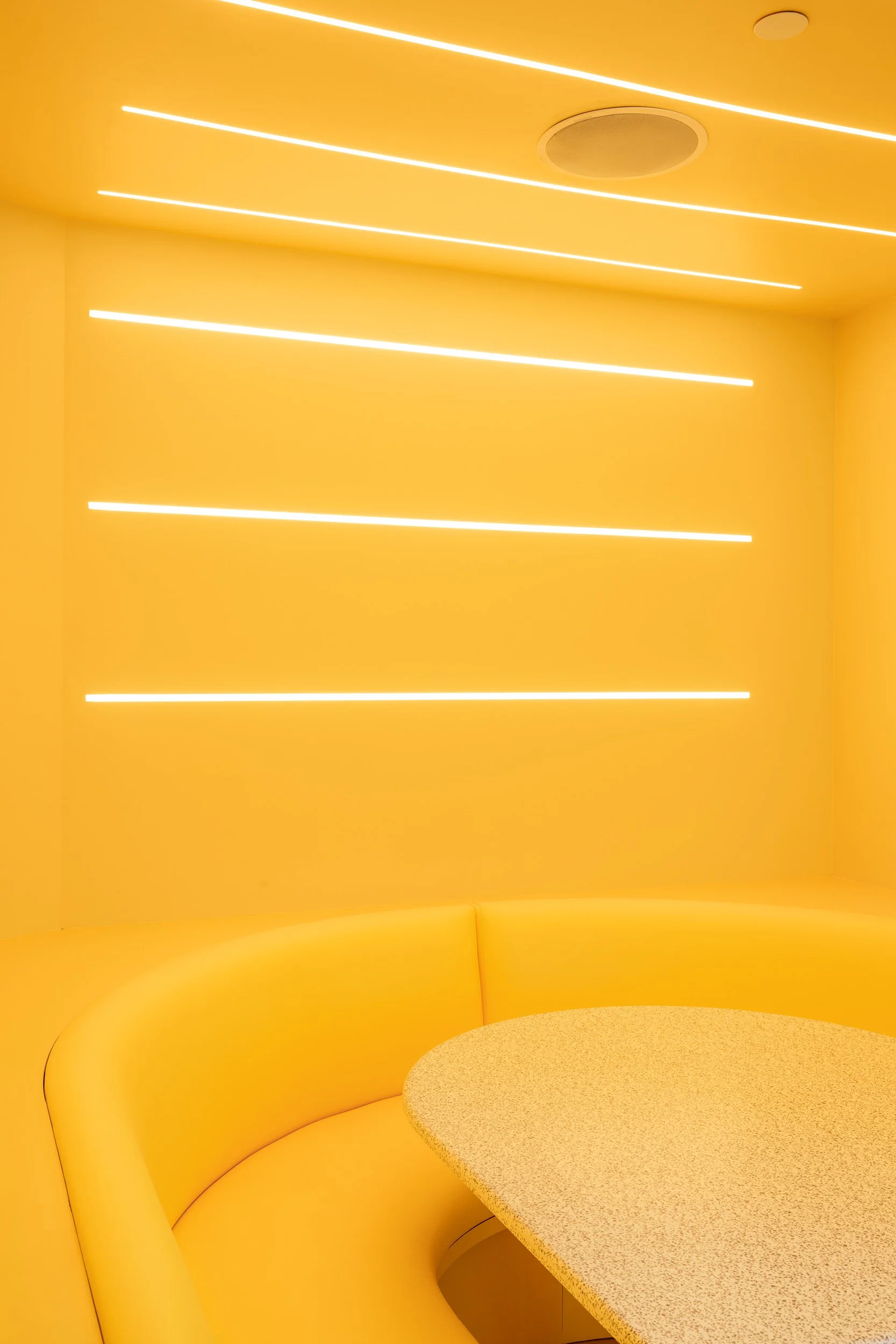A bright yellow room with curved yellow seating and a yellow speckled table. The wall features horizontal white LED light strips.
