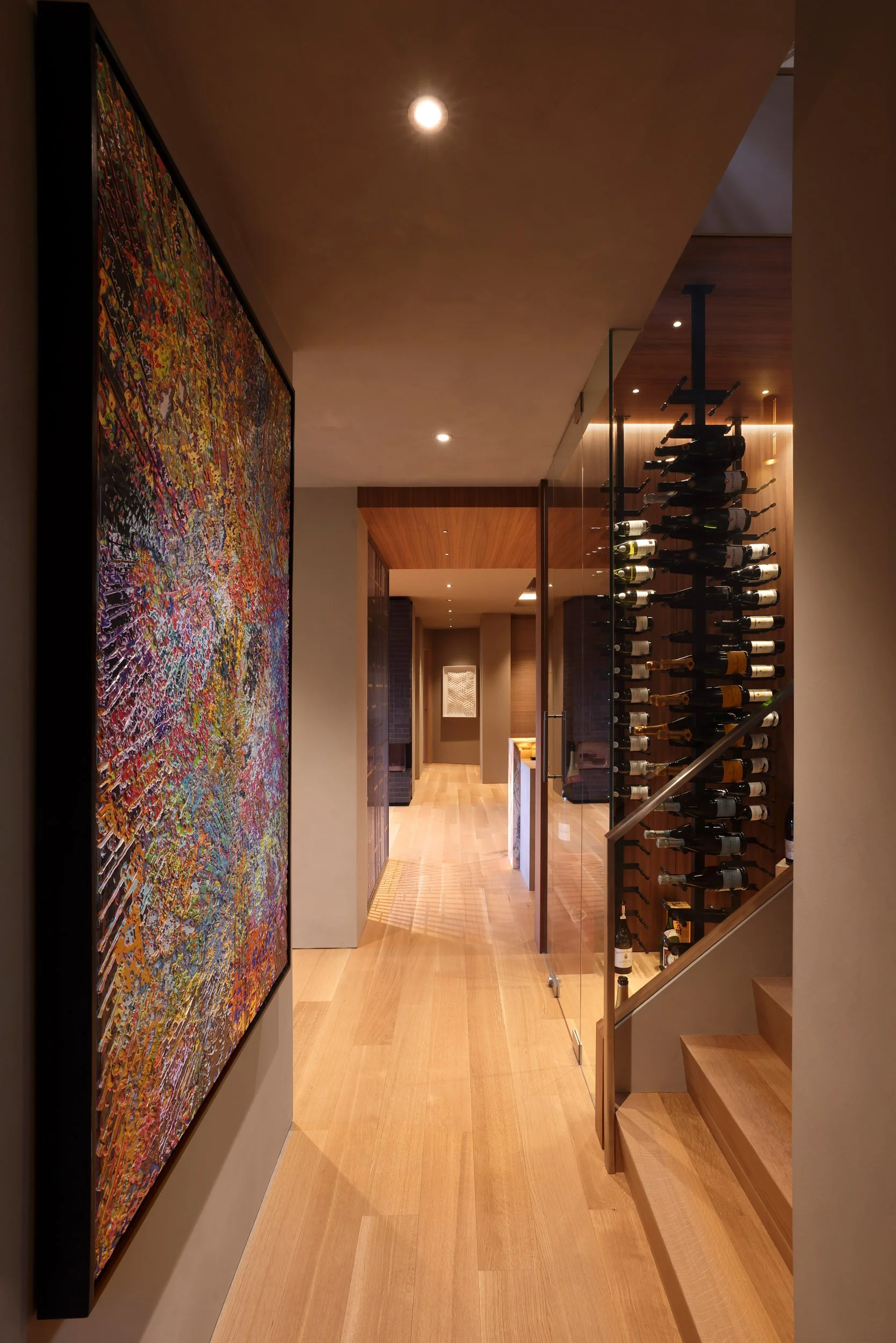A hallway with wooden flooring, artwork on the left wall, a glass door on the right, and a wine rack visible through the glass.