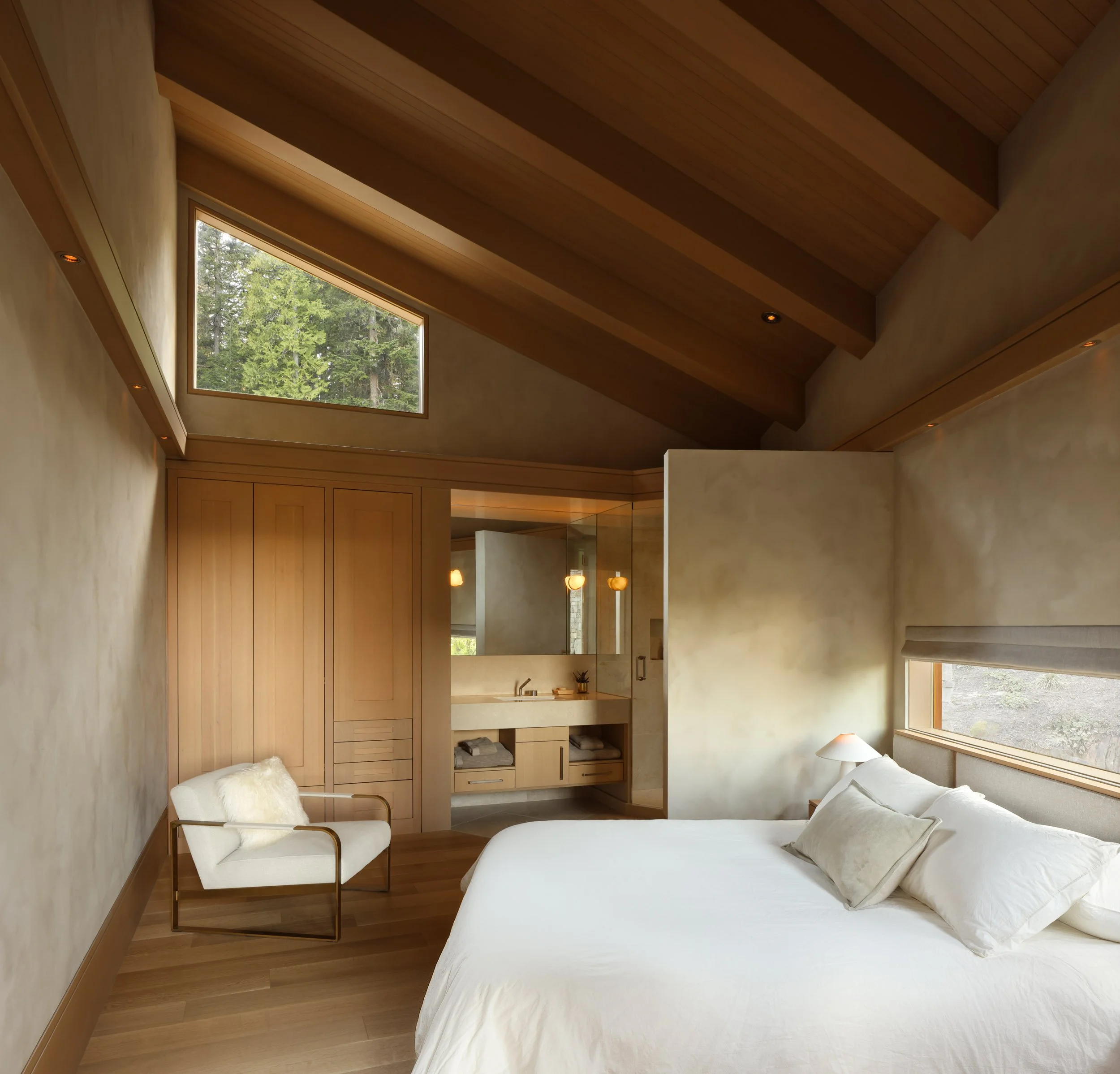 A bedroom with a vaulted wooden ceiling, a large window showing trees outside, a white bed with pillows, a bedside lamp, a white armchair with a furry pillow, and a view of a bathroom with a mirror and sink.