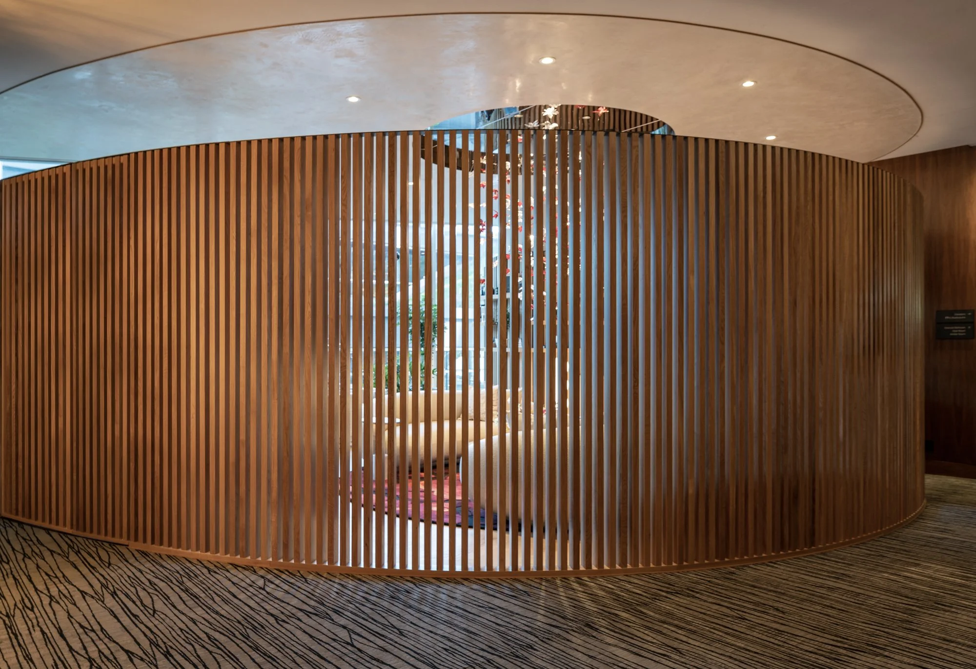 Interior view of a modern space with a wooden slatted curved wall and a seating area visible through the slats, with a ceiling featuring recessed lighting and a patterned carpet.