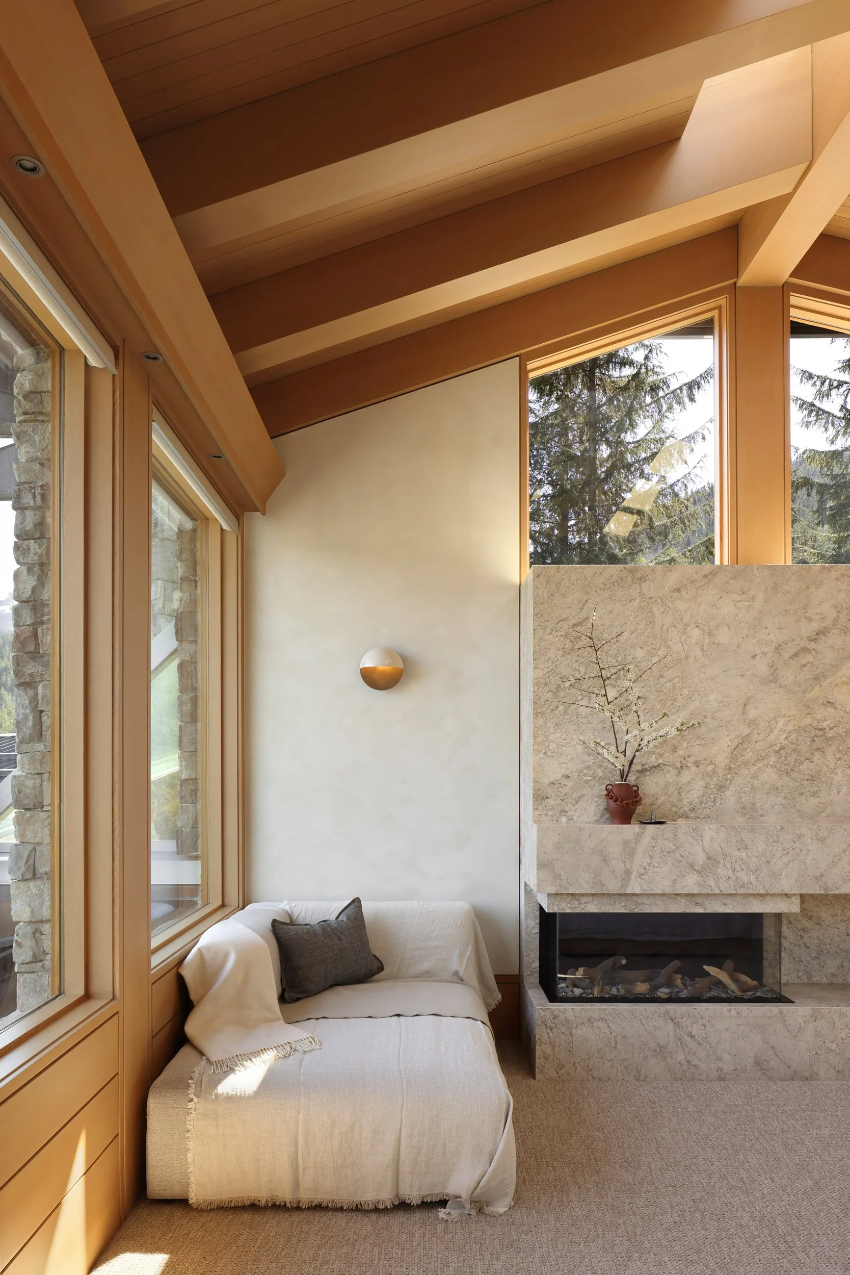 Living room corner with beige sofa, dark gray pillow, large windows, wooden ceiling beams, stone fireplace with decorative vase and branches, and a carpeted floor.