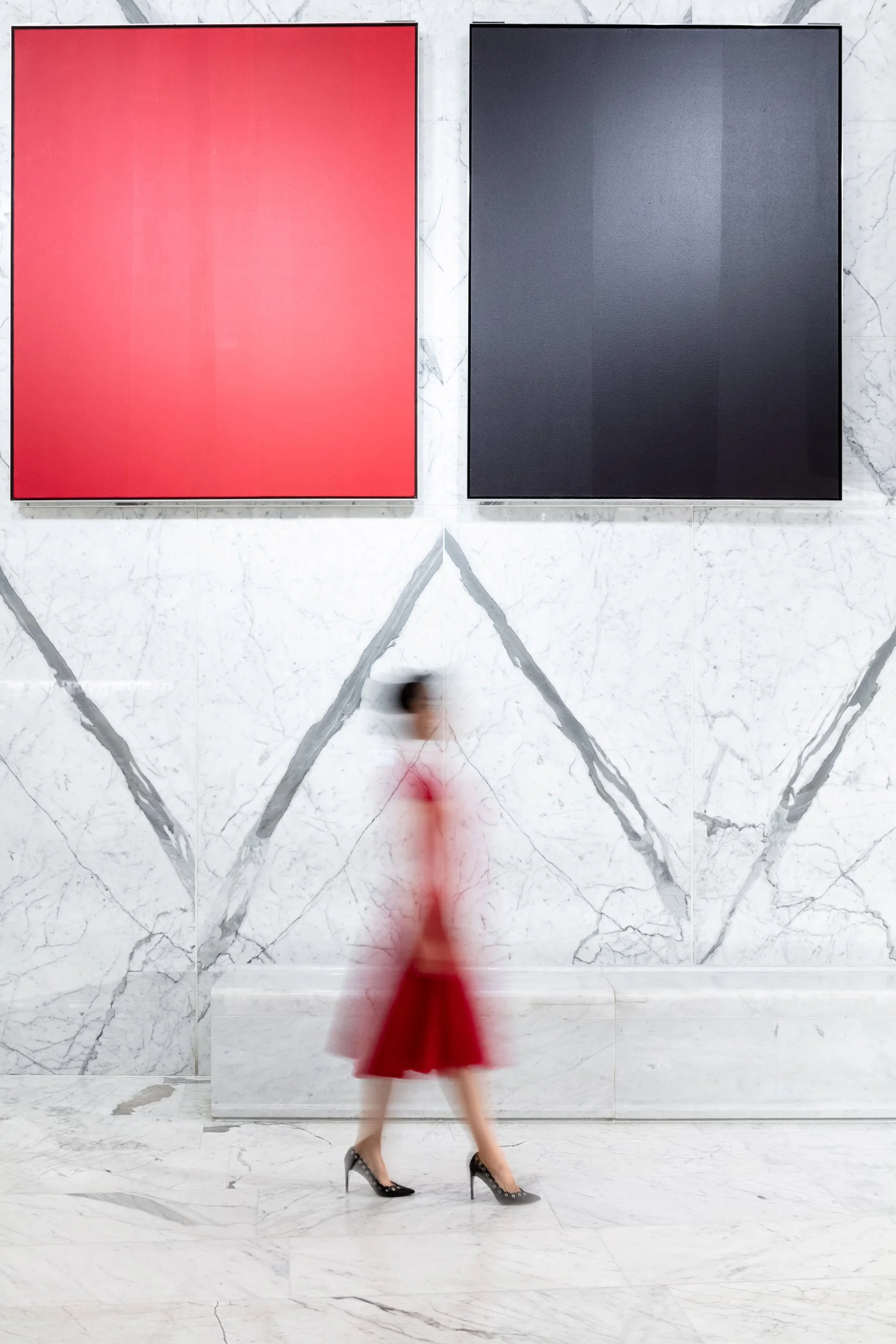 A woman in a red dress and high heels walking past two large, framed art pieces on a white marble wall in an art gallery.