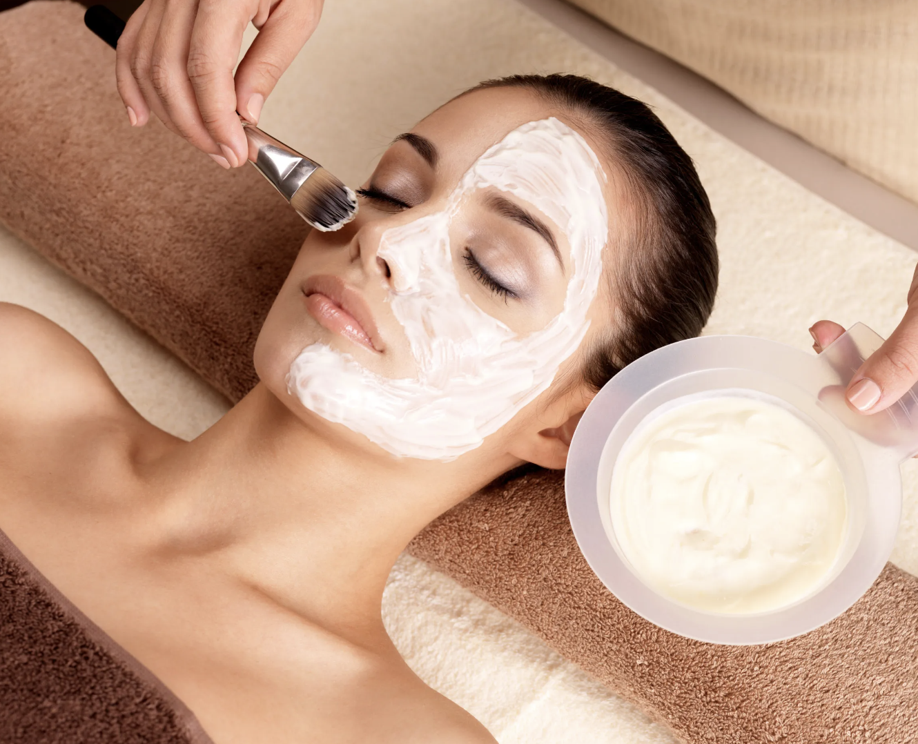Person receiving a facial treatment with a white cream mask applied with a brush.