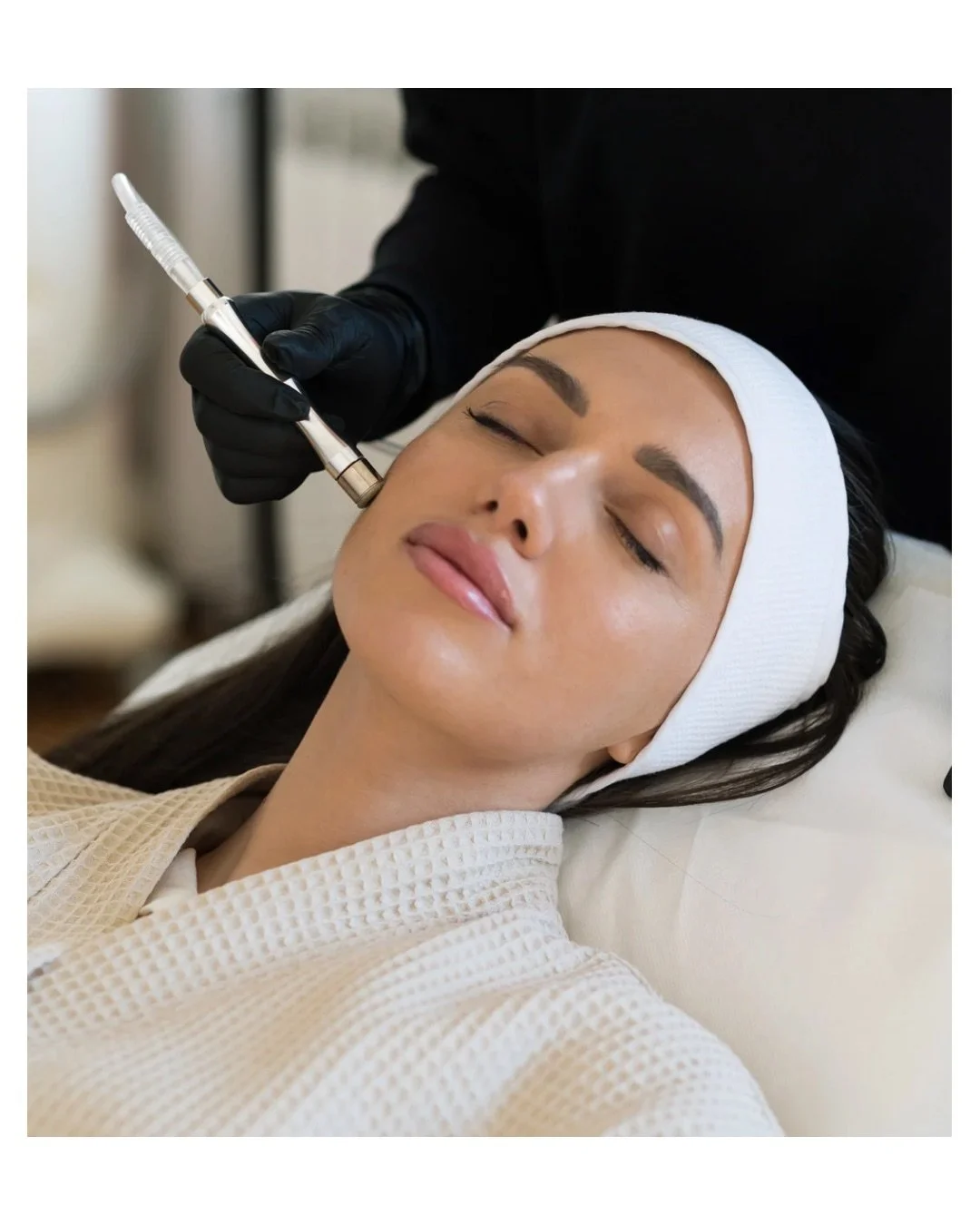 Woman receiving a cosmetic facial treatment with a syringe by a professional wearing black gloves, lying on a treatment bed with her eyes closed, wearing a white headband and robe.